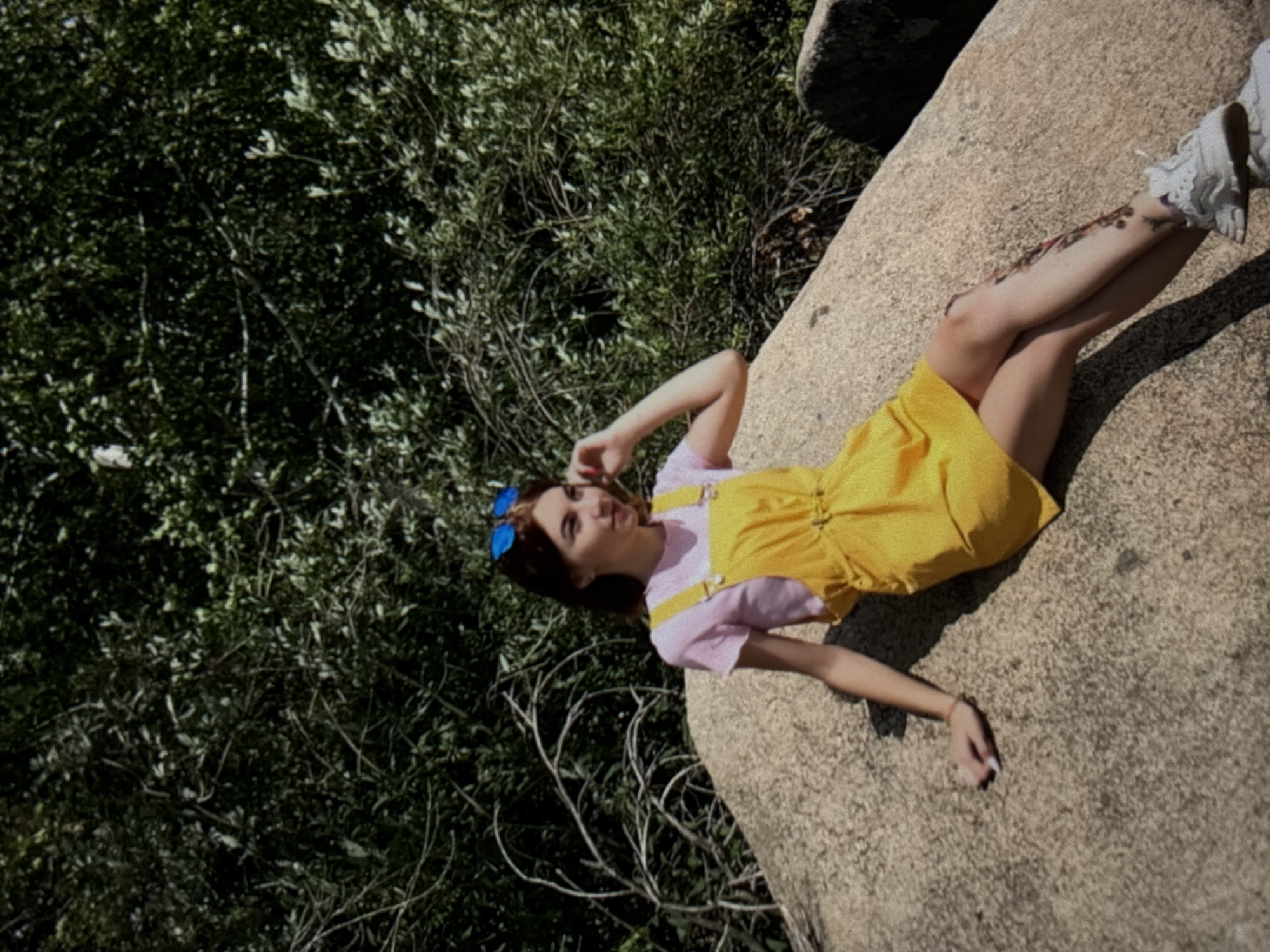 A young woman with auburn hair, wearing a pink t-shirt, yellow overalls, blue sunglasses, and white sneakers, is sitting on a large rock. She is touching her hair with her right hand and her left hand rests on the rock. A tattoo is visible on her right leg. The background is filled with dense green foliage and dry branches. The scene appears to be outdoors, possibly in a park or a natural area, under a bright sky. There is no discernible Omsk-specific detail in the image.