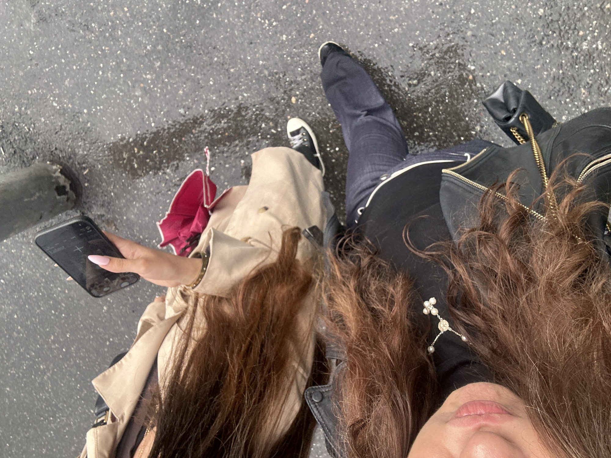 An overhead view captures two individuals standing on wet, dark paved ground, likely a street or sidewalk in Yerevan, Armenia. The damp surface suggests recent rain or drizzle.
On the left, a person with long, straight brown hair wears a light beige trench coat. Their hand, adorned with light pink nail polish, holds a black smartphone displaying a faint "+" icon on its screen. A partially visible pink umbrella is tucked beside them. They are wearing black sneakers with white soles.
On the right, another person with long, curly brown hair is visible. They wear a black top or jacket, featuring a unique pearl and silver accessory. Their lower lip and chin are seen looking upwards, suggesting they are holding the camera. This individual wears dark blue jeans and black sneakers. A black handbag with a gold chain strap is also visible near their side. The overall mood appears casual, with a hint of a smile on the visible lips of the person on the right.