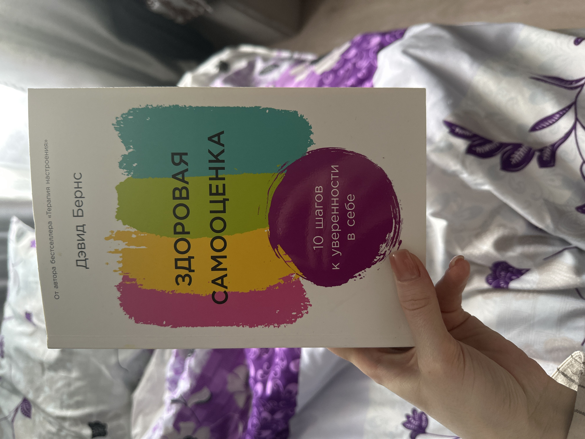 A person is holding a book titled "Здоровая Самооценка" (Healthy Self-Esteem) by David Burns, in Russian. The book's cover features a design with horizontal brushstrokes in teal, lime, magenta and a purple circle containing the text "10 шагов к уверенности в себе" (10 Steps to Confidence in Yourself). The book is held against a background of crumpled bedding with a white base and purple floral patterns. The location, Pokrov, Russia, is not ascertainable from the image.