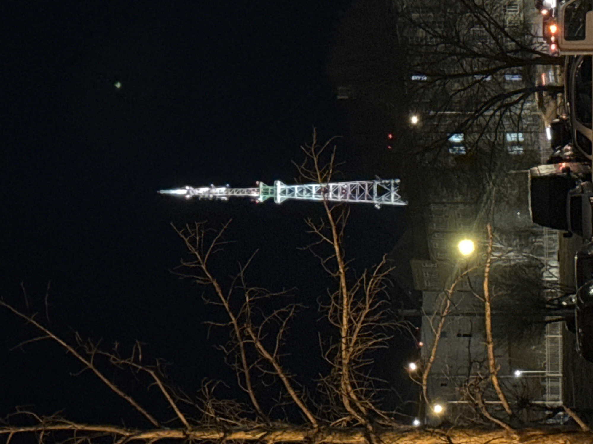 The image shows a nighttime view of a tower in Krasnoyarsk, Russia. The tower is illuminated with colored lights. In the foreground are bare tree branches. In the background, there are buildings, street lights, and vehicles.