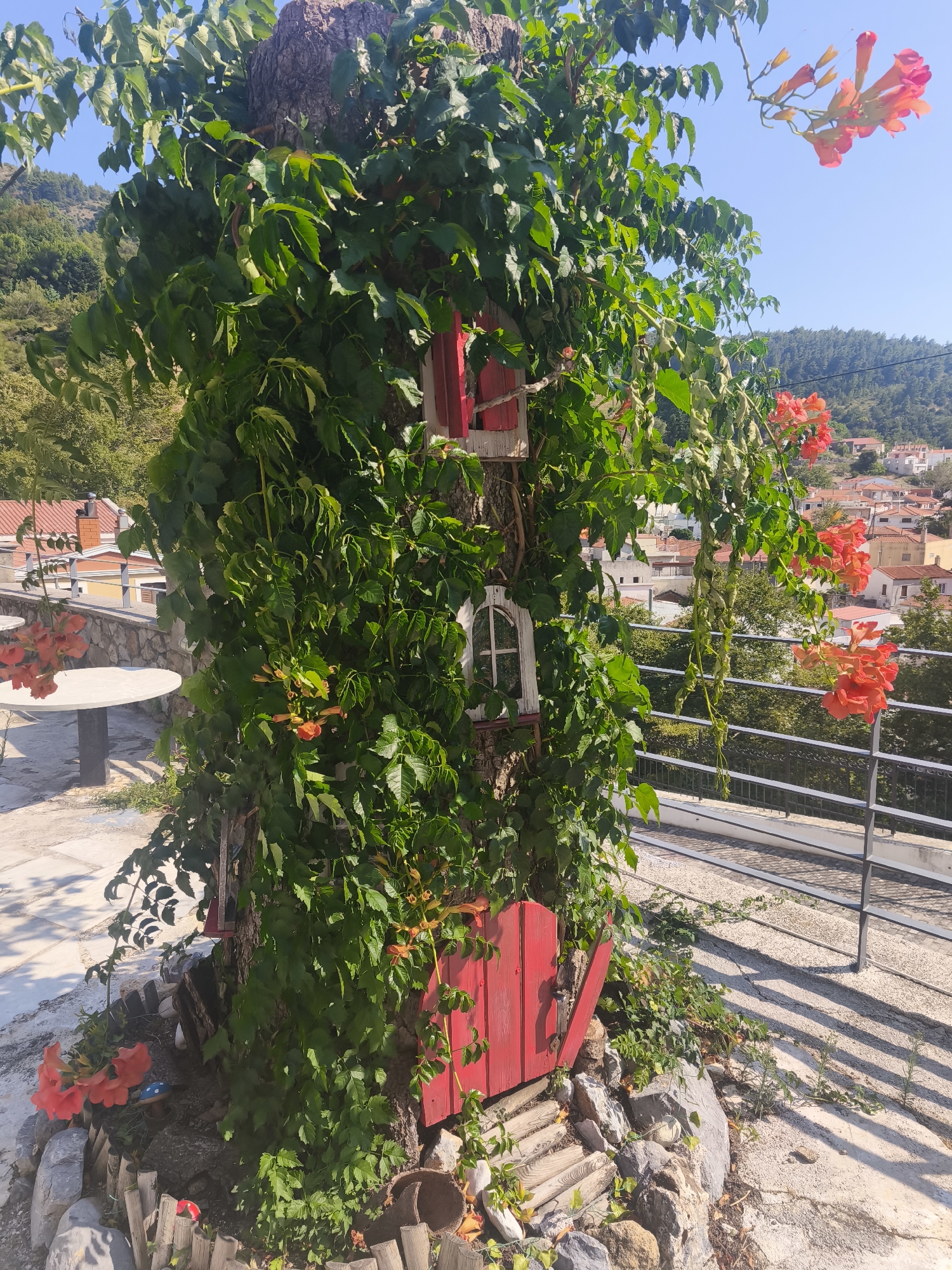 In the city of Stené Dirfyos, Greece, a large tree trunk is adorned with lush green foliage and vibrant orange trumpet flowers. Several miniature red doors and windows are integrated into the tree's base, giving it the appearance of a whimsical dwelling. The surrounding area features a paved patio with a white circular table and chairs, a stone wall, and a metal railing overlooking a village with red-tiled roofs nestled amongst rolling hills. The sky is clear and blue, indicating a sunny day.