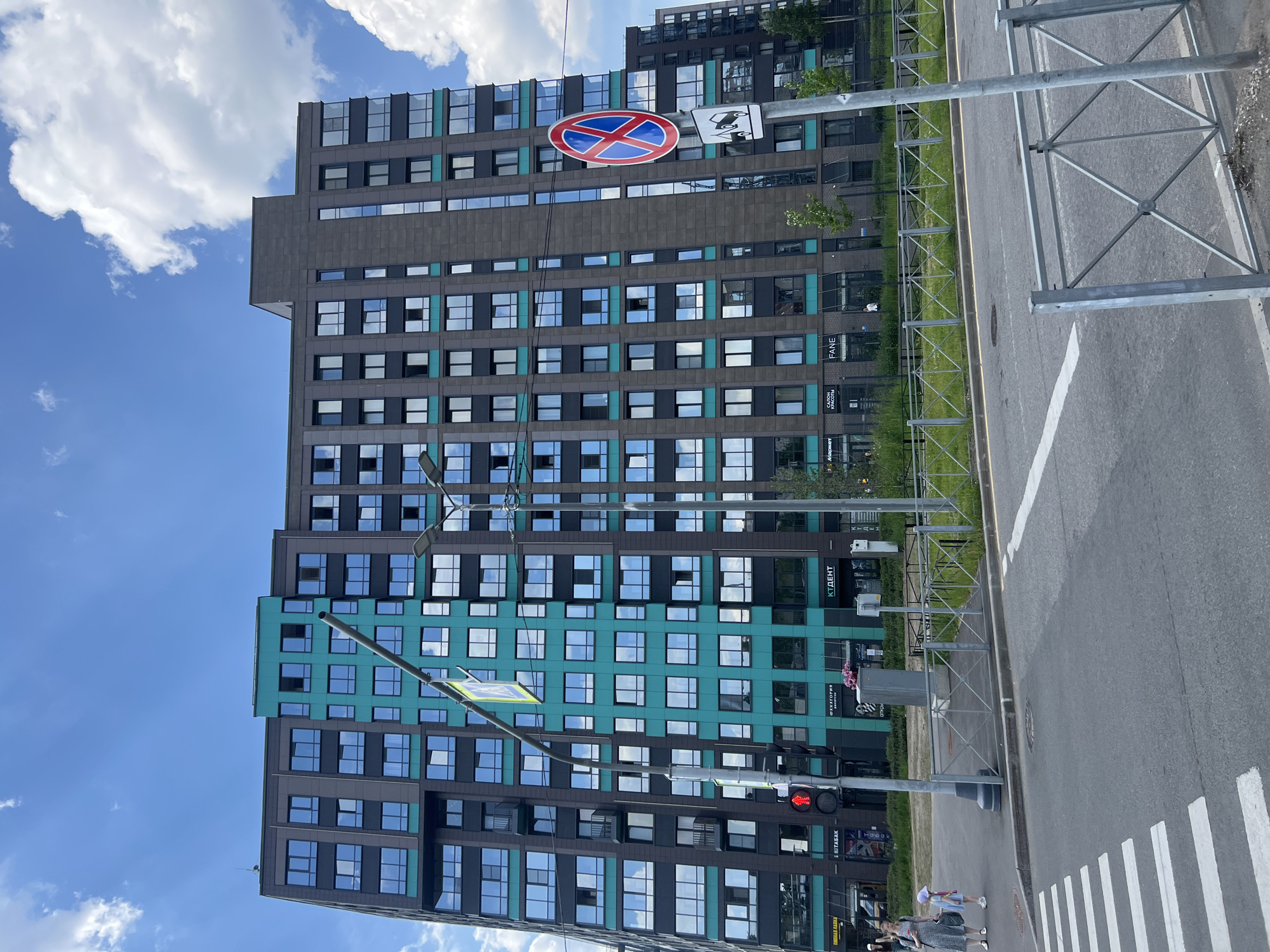 The image depicts a modern urban street scene in Saint Petersburg, Russia, under a bright blue sky with scattered white clouds. The primary subject is a contemporary multi-story building featuring a striking facade of dark gray/brown and teal/turquoise panels, punctuated by numerous reflective rectangular windows.
A paved road with a distinct white-striped crosswalk runs alongside the building. Separating the road from the structure is a grassy strip, bordered by a metal fence. Prominently displayed overhead are road signs: a circular red sign with a blue cross, indicating "No Stopping" or "No Parking," and an adjacent white rectangular sign with vehicle icons. A traffic light, showing a red signal, is also visible near the crosswalk.
On the sidewalk, near the crosswalk, two individuals—an adult woman in a patterned dress and a child in light clothing—are seen walking away from the camera. The time of day is clearly daylight, indicated by the bright sky and strong reflections in the building's windows.
Several signs with Cyrillic text are visible on the building, confirming the Russian location. These include "ФАНЕ" (Fane), "САЛОН КРАСОТЫ" (Beauty Salon), "МАРКЕТ" (Market), "КТДЕНТ" (KT DENT), "ЭЛЕКТРОНИКА" (Electronics), "5 ЭТАЖ" (5th Floor), and "ШВАРМА" (Shavarma), suggesting a mix of commercial and possibly residential spaces. The scene captures a typical, unhurried moment in a modern urban environment.