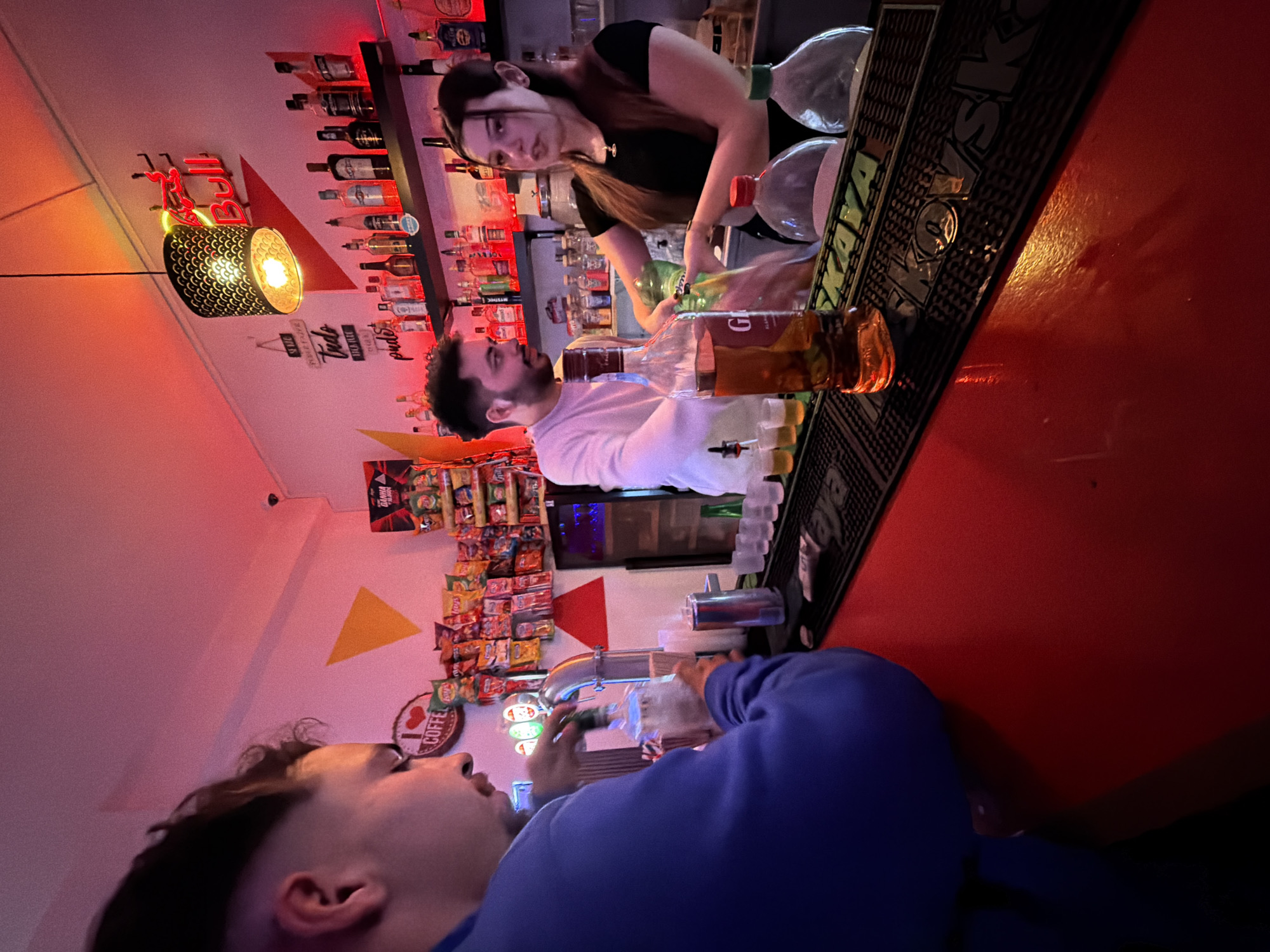 The image depicts an indoor scene at a bar in Vila Real, Portugal. A male customer in a blue sweatshirt is sitting at the bar, looking up. Behind the bar, a male bartender in a white shirt and a female bartender in a black shirt are preparing drinks. Various bottles of alcohol are displayed on shelves above the bar, with a Red Bull sign visible on the left. There are also snacks and a "coffee" sign visible near the customer. The bar counter is red with "KAYAKA" engraved on it. The lighting is somewhat dim, with red and yellow tones creating a casual atmosphere.