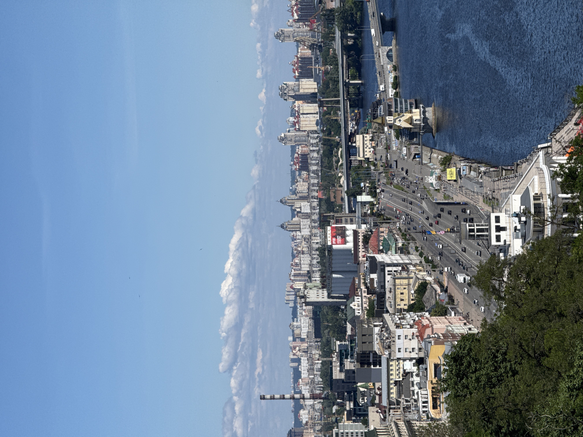 This is a high-angle, full-shot, eye-level, long shot photograph taken from a distance, capturing a panoramic view of the city of Kyiv, Ukraine, on a clear, sunny day. The image features a wide, blue river flowing from the right to the left, with several bridges crossing it. The foreground is dominated by lush green trees on the left, and a white, modern building with curved balconies on the lower right.
In the mid-ground, a bustling multi-lane road runs parallel to the river, with numerous cars and trucks visible. A distinctive, whimsical building shaped like a golden clock tower stands prominently on the riverbank. Beyond the road and river, the cityscape unfolds with a dense collection of buildings of varying heights and architectural styles. Tall, modern apartment buildings with light-colored facades and reddish roofs are clustered together, interspersed with industrial structures, including a prominent red-and-white striped smokestack on the left. Cranes are visible in the distance, suggesting ongoing construction or industrial activity. The sky is a vibrant blue, with scattered white clouds. A few birds are faintly visible in the sky.