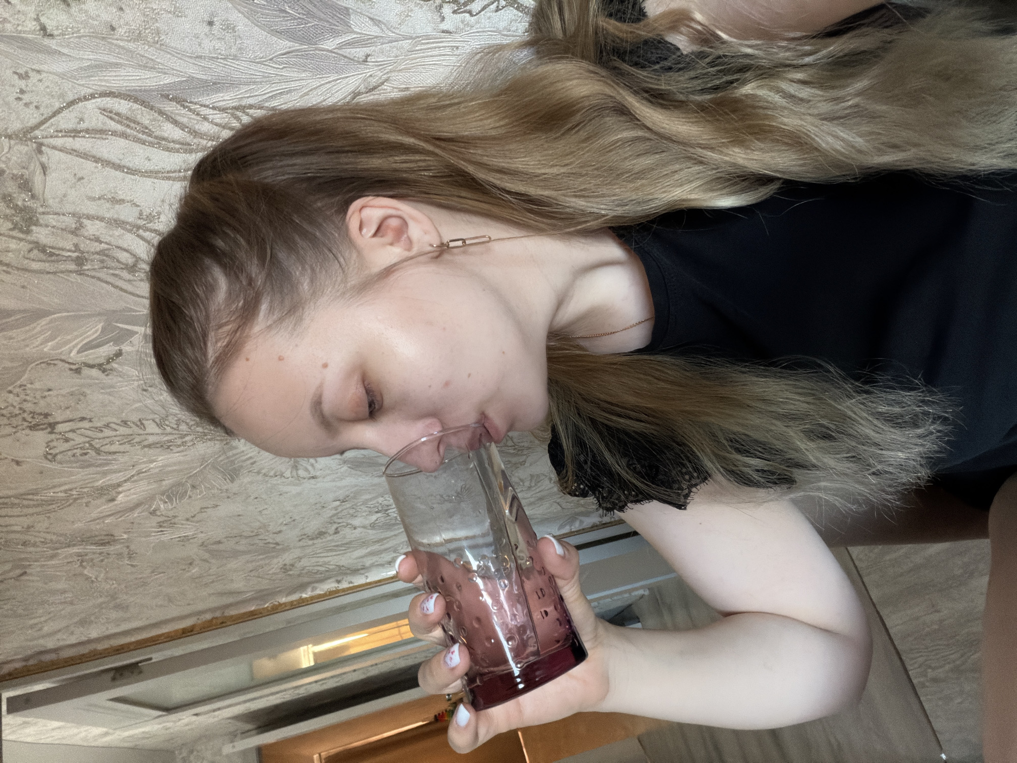 An indoor scene in the city of Reshetnikovo, Russia, shows a young adult female in profile, facing left, drinking from a glass. She has long, wavy, blonde hair, partially styled in a high ponytail with loose strands. Her complexion is fair with visible facial blemishes. She wears a black top featuring black lace trim on the shoulder, a thin gold-colored necklace chain, and a gold-colored rectangular link dangle earring on her right ear. Her right hand, adorned with white nail polish featuring small red and white designs, holds a clear glass with a textured, purple-tinted base and raised circular patterns, containing a translucent liquid. The background wall is covered in light-toned wallpaper with a subtle, reflective leaf-like pattern. To the left, a light-framed doorway or mirror reflects an interior area with warm lighting. A light wood-effect floor is visible in the lower left foreground.