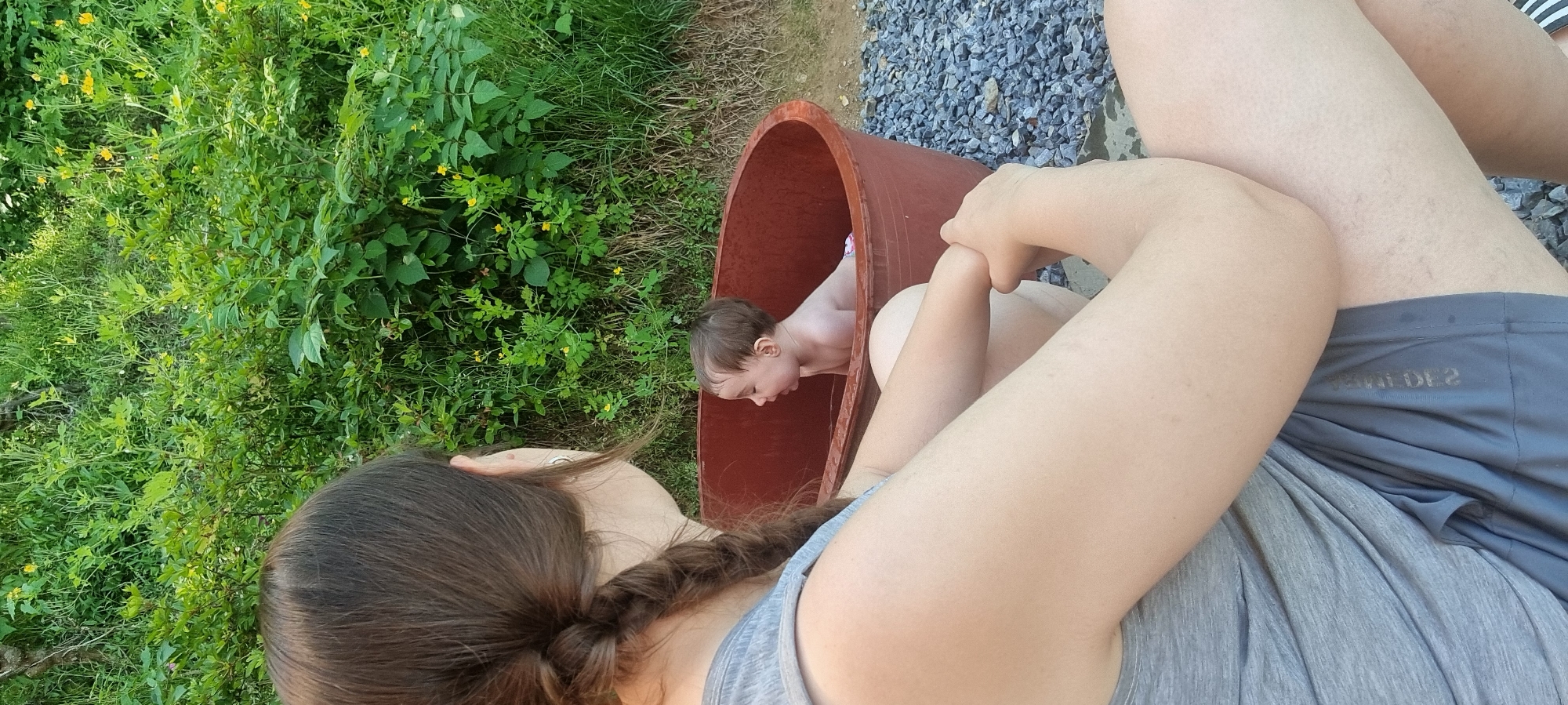 The image shows a baby bathing in a brown, oval-shaped tub. A person with long, braided brown hair sits beside the tub. Next to the tub are gravel, and a person wearing gray shorts and a gray shirt. Green plants are visible in the background. The image was taken outside, likely in Dunnae-myeon, South Korea.