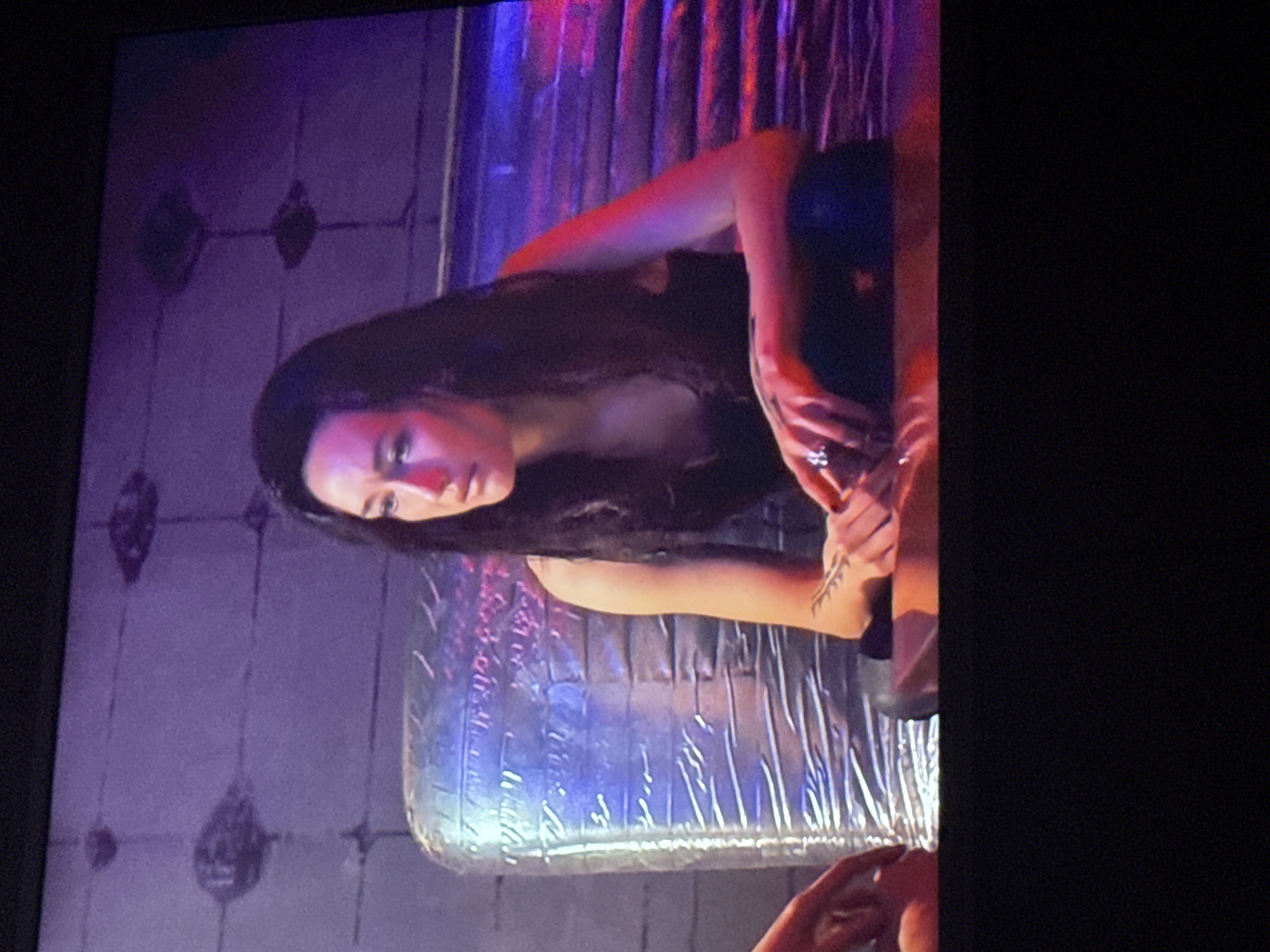 A woman with long, dark hair is seated on a bed that has silver-colored plastic sheeting covering it. She is wearing a dark-colored top or dress and has a ring on her finger. Her left arm, which has a tattoo on the inner forearm, is visible. The background consists of a wall with a light purple hue and a pattern of dark, oval-shaped designs. The image appears to be taken from a slightly upward angle, possibly from a television screen. There is no information in the image or available to me regarding the location being Mytishchi, Russia.