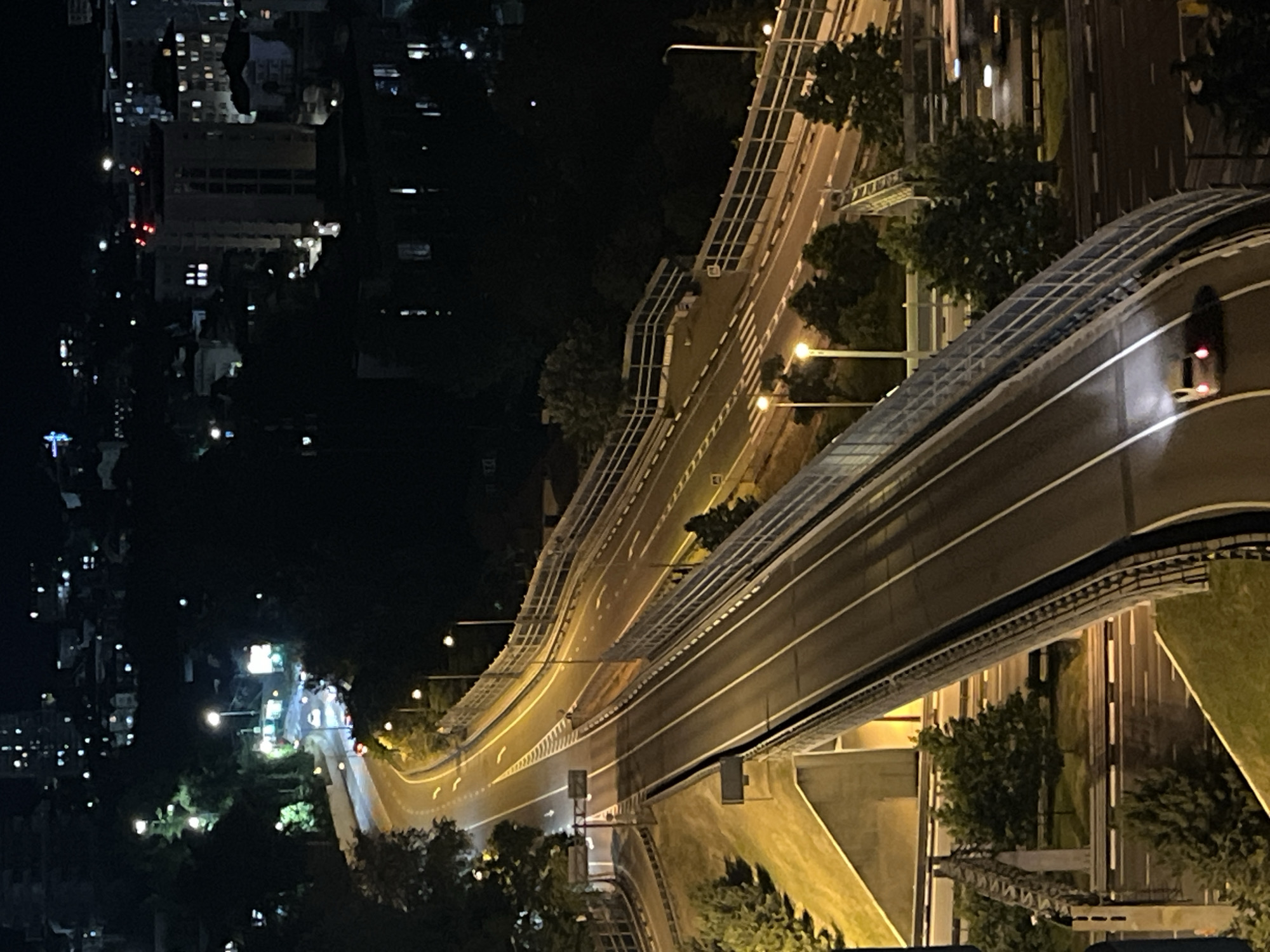 The image shows an elevated highway at night, with city lights and buildings in the background. The highway is illuminated by streetlights and the headlights of a few cars. The foreground features a single car with its taillights illuminated, driving on the highway. The environment appears to be a hilly or mountainous area, consistent with the topography of Sochi, Russia. The city in the background is densely populated with buildings of various heights. The overall impression is one of urban infrastructure at night, with a sense of depth and scale provided by the elevated highway and the distant cityscape.