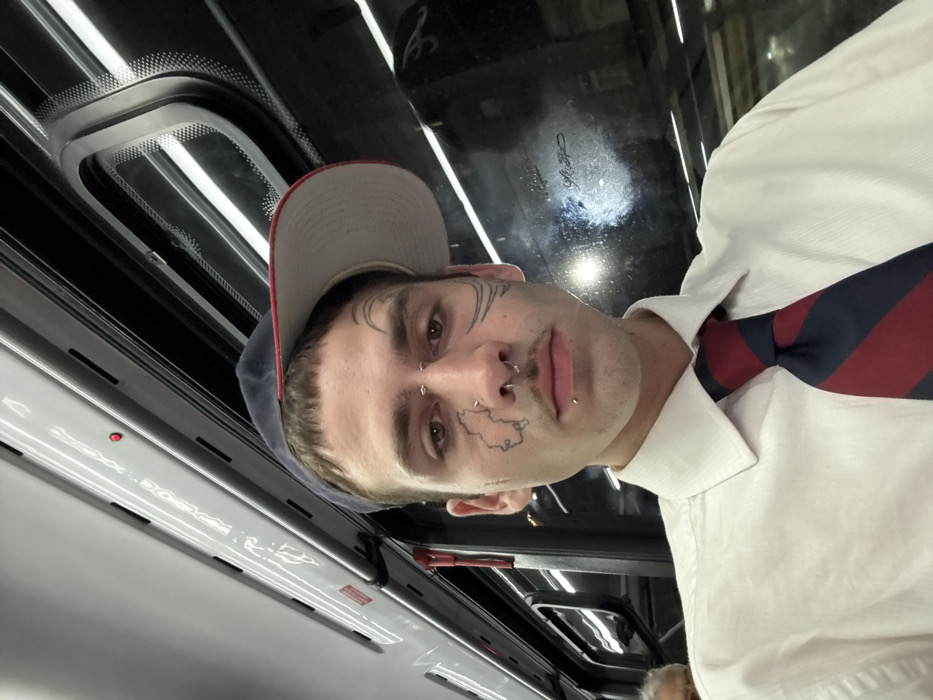 The image depicts a young adult male inside a subway train (Subte) in Buenos Aires, Argentina. The male subject, positioned centrally, has light skin, short brown hair with bangs, and a thin mustache. He is wearing a white collared shirt, a dark blue and red striped necktie, and a navy blue baseball cap with a light-colored brim worn backward. His face features multiple piercings: a septum ring, two nostril rings, a vertical barbell below his lower lip, and two horizontal barbells above his right eyebrow. Distinct tattoos are visible on his face, including a stylized map outline on his left cheek, abstract curved lines on his right temple/cheekbone area, and a small crescent moon above his left eyebrow. He is looking directly forward, seemingly at the camera. In the lower-left corner, a portion of an older individual's head with grey hair is partially visible, indicating another passenger. The environment consists of the train's interior with a grey ceiling, linear fluorescent lighting, and black-framed windows. The window on the right shows reflections of the interior lights and a dark, blurred exterior, consistent with a moving train, possibly within a tunnel or during nighttime.