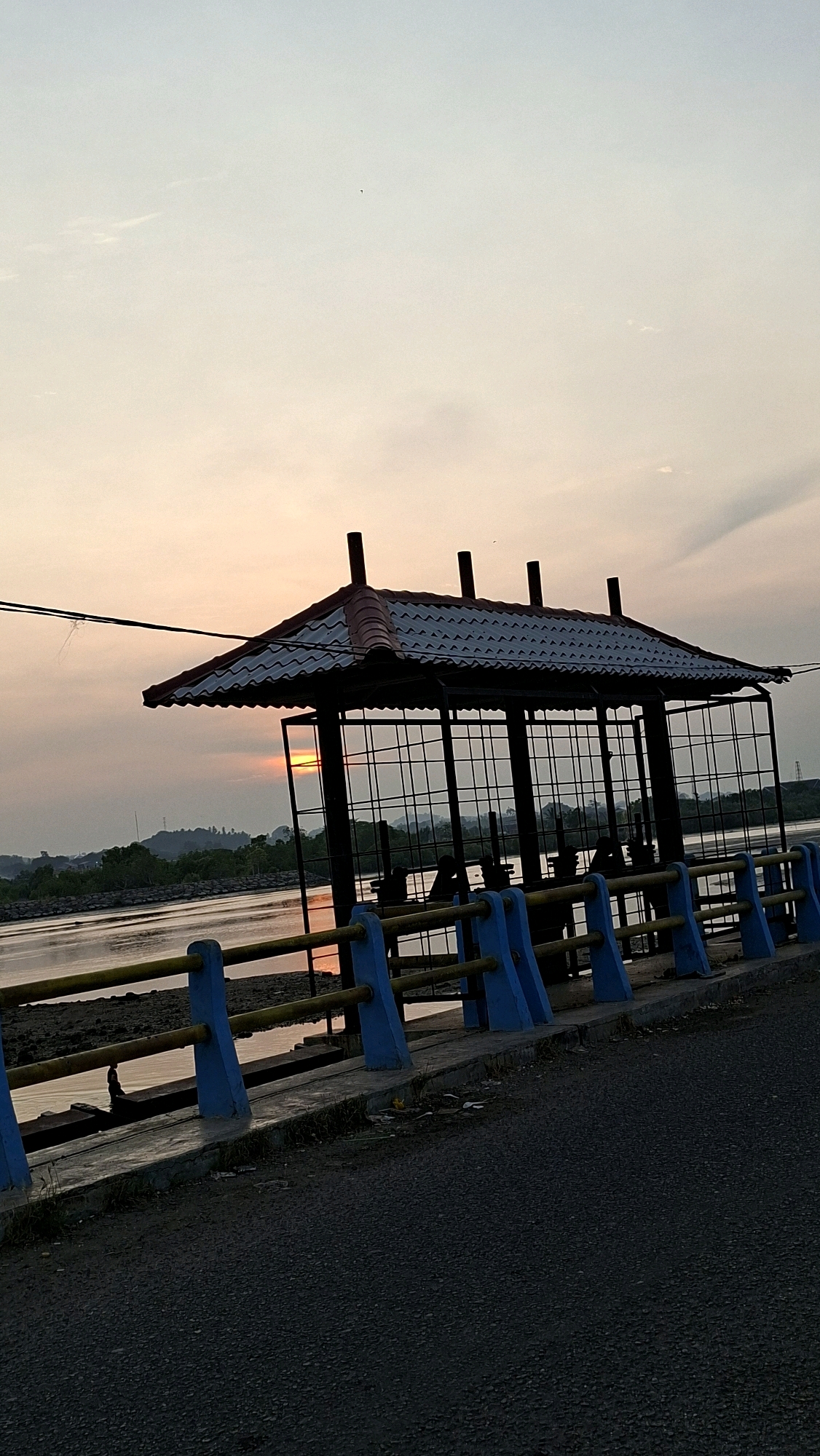 The image shows a gazebo structure located on a causeway next to a body of water. The gazebo has a tiled roof and metal bars forming its enclosure. A fence with blue supports and yellow railings runs along the edge of the causeway. In the background, there are trees and hills, and the sun is setting over the horizon, casting a warm glow. The water reflects the colors of the sunset. The causeway is paved with asphalt. Silhouettes of people can be seen inside the gazebo. The scene appears to be a waterfront area in Lhokseumawe, Indonesia, possibly a recreational spot.