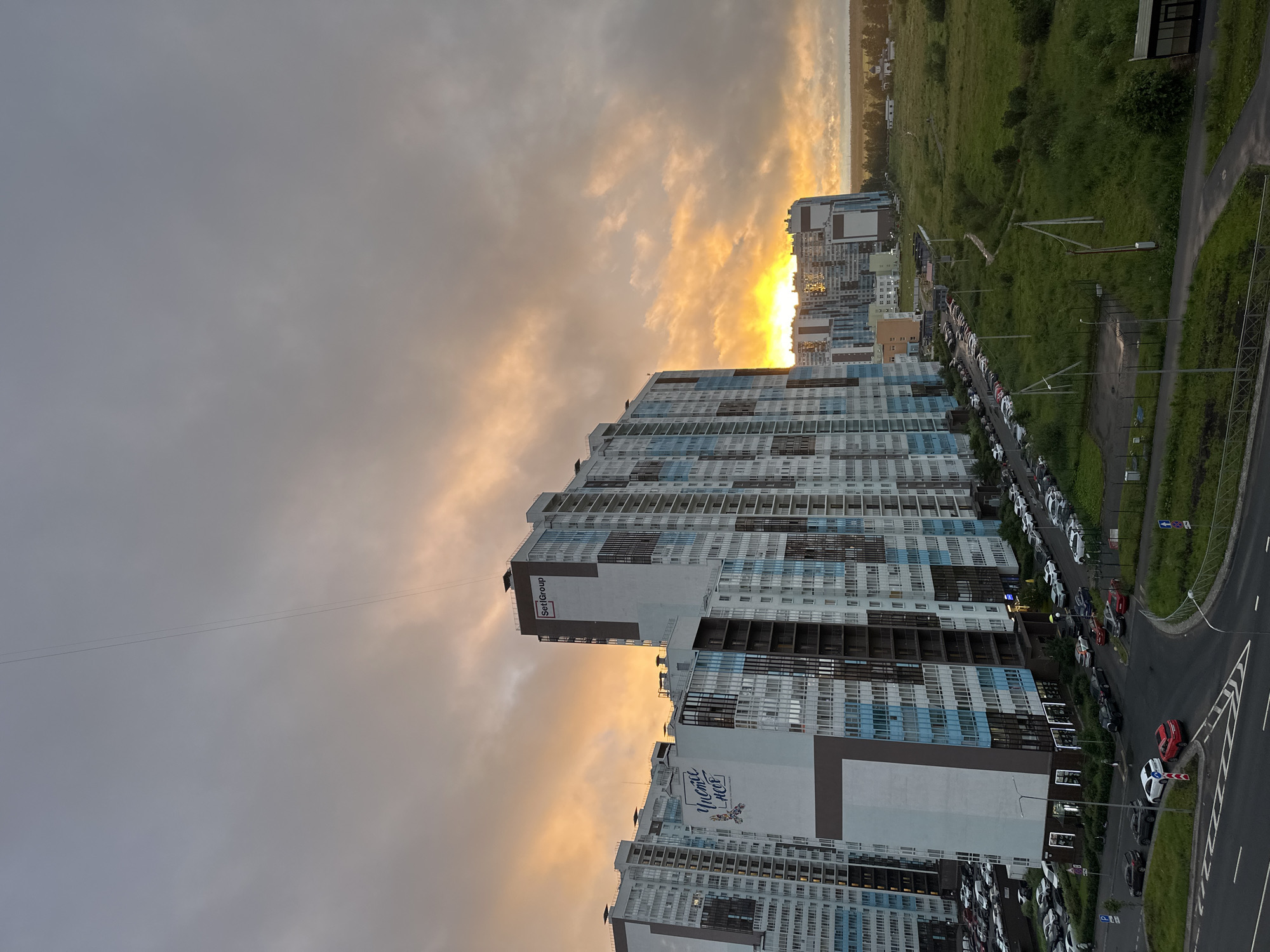 The image depicts a panoramic view of a residential complex in Saint Petersburg, Russia, during a sunset. The sky is filled with dramatic, cloudy formations tinged with hues of orange and yellow, indicating the sun is setting or has just set. Several modern, multi-story apartment buildings dominate the mid-ground, characterized by their white, grey, and blue facades. One building prominently displays the "SetIGroup" logo.
In the foreground, a street is visible with numerous cars parked along the side and driving on the road. The architecture suggests a relatively new development. To the right of the buildings, a vast expanse of green, uncultivated land stretches towards the horizon, dotted with trees and a small building that appears to be a church or chapel. The overall atmosphere is one of urban development encroaching on a natural landscape, set against a vibrant, cloudy sky. The image is taken from an elevated perspective, likely from a window or balcony of another building.