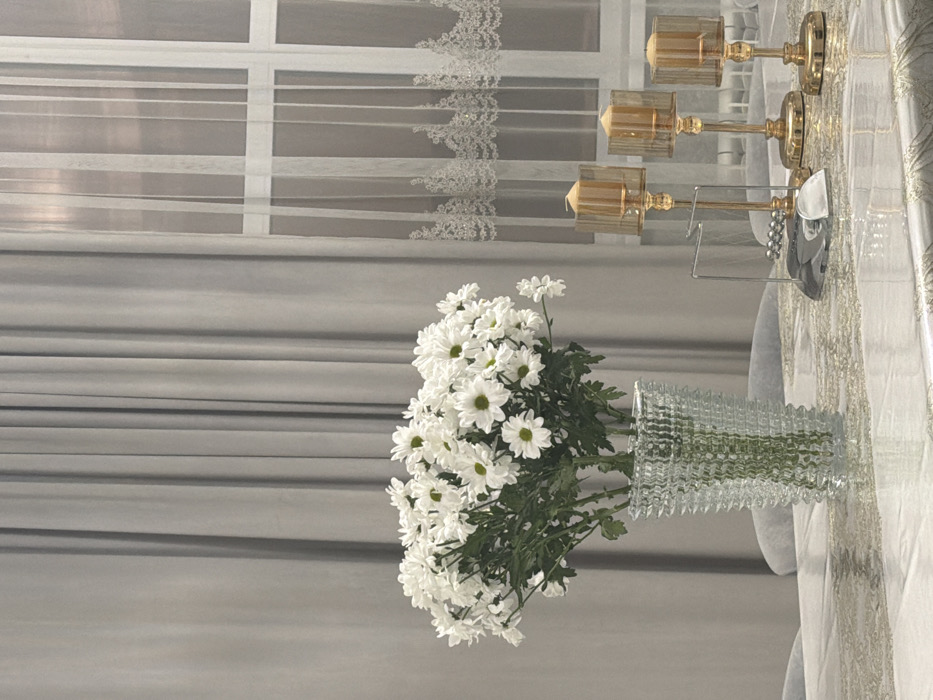 White daisies fill a clear, textured vase, placed on a table next to decorative golden candelabras. The table is covered with a white tablecloth adorned with gold accents. The background features a gray curtain and a white window curtain with lace trim.