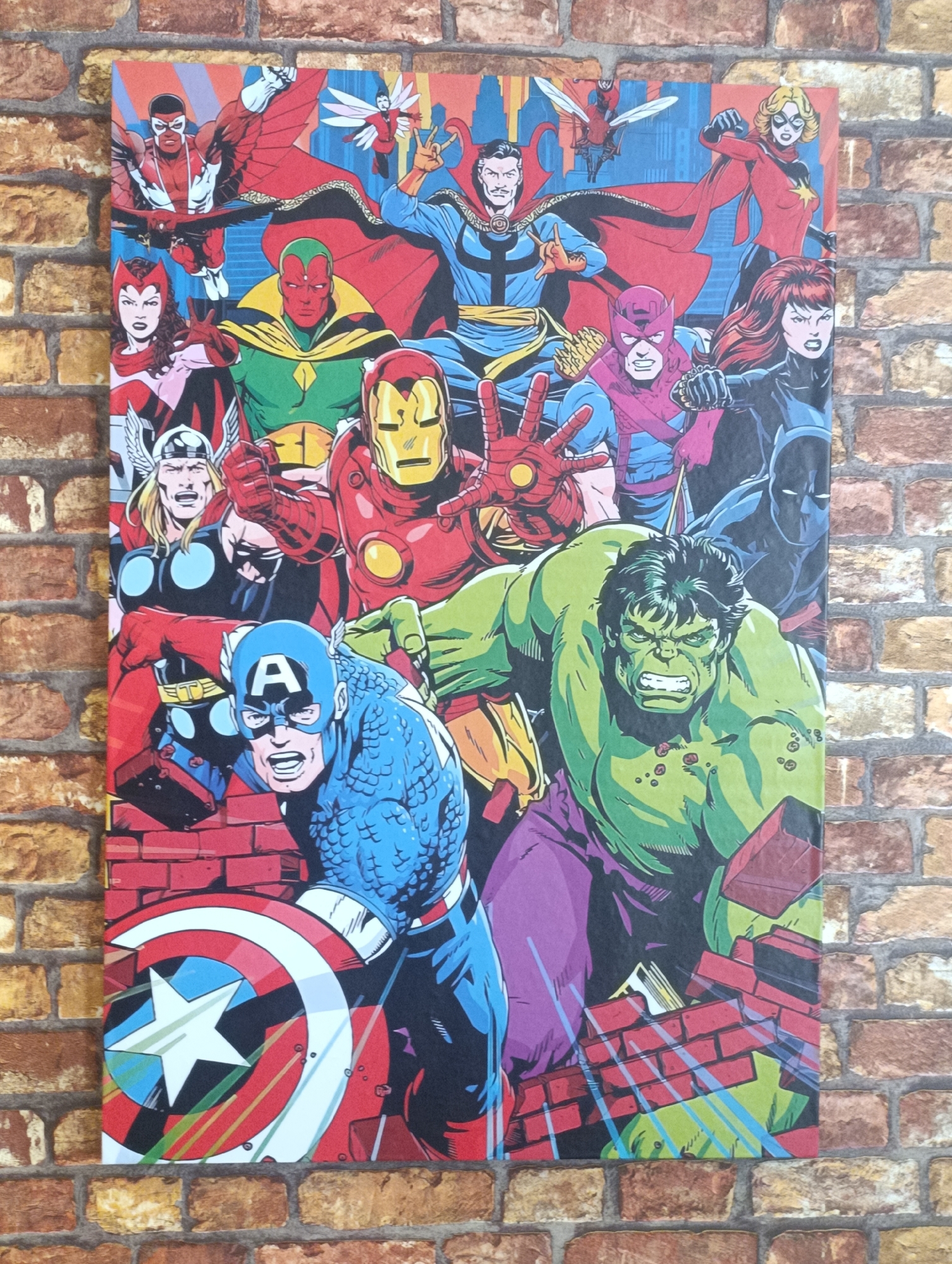 The image is a photograph of an Avengers comic book print hanging on a brick wall. The comic book print features several Marvel superheroes, including Captain America, Hulk, Iron Man, Thor, Vision, Scarlet Witch, Black Panther, Doctor Strange, Hawkeye, Black Widow, Falcon, Ant-Man, and Ms. Marvel. They are posed together in a dynamic group shot. The bottom of the image shows remnants of brick structures, suggesting the superheroes have broken through them. The wall is made of reddish-brown bricks with light brown grout. The location is not provided in the image.