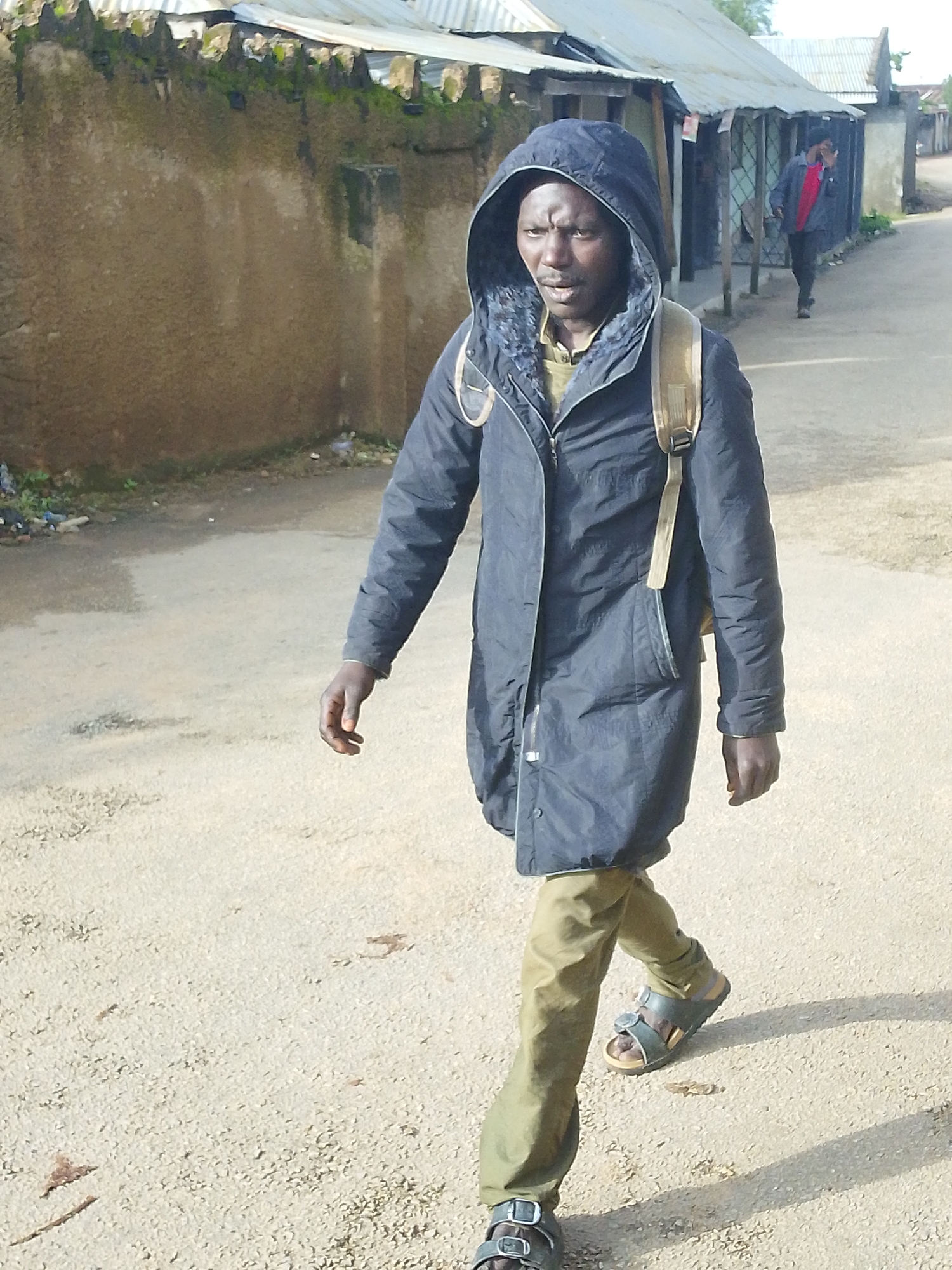 The image depicts a man walking on a dirt road in Jos, Nigeria. He is wearing a dark hooded jacket, khaki pants, and sandals. He is also carrying a backpack. In the background, there are buildings with corrugated metal roofs and a second man in a red shirt walking away. The ground is a mixture of dirt and gravel, with some scattered debris. The wall on the left is rough and appears to be made of concrete or mud. The overall scene suggests a somewhat impoverished urban environment.