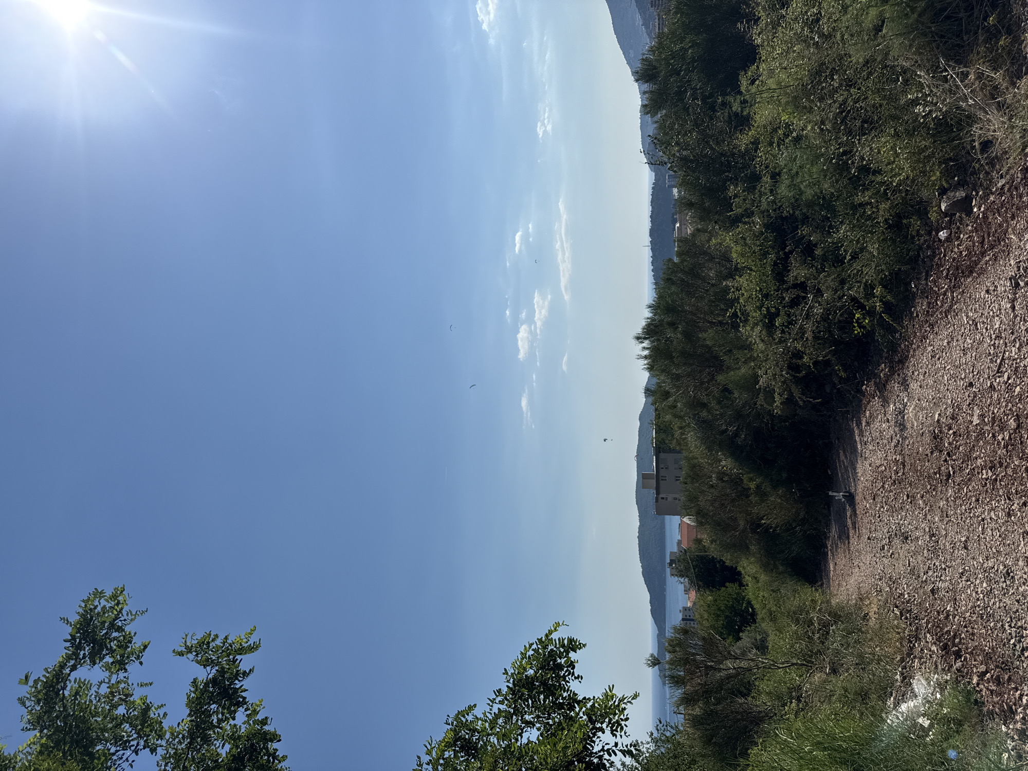 The image depicts a bright, sunny day in Rafailovići, Montenegro, with a clear blue sky and scattered white clouds. The sun is prominently visible in the upper left corner, casting a lens flare. The scene is dominated by lush green vegetation, with trees and shrubs lining a rough, gravel path that winds through the foreground and midground.
In the distance, a body of water, likely the Adriatic Sea, is visible, bordered by a hilly coastline dotted with buildings. The architecture suggests a coastal town, with some structures appearing to be residential or possibly hotels. The hazy atmosphere indicates it's a warm day. There are a few small, dark shapes in the sky, which could be birds or perhaps paragliders, adding a dynamic element to the otherwise static landscape.
The composition is oriented vertically, giving a sense of looking up towards the sky and across the landscape. The contrast between the vibrant green foliage and the stark gravel path is striking. The overall mood is serene and suggests a peaceful, natural environment with hints of human habitation. No text is visible in the image.