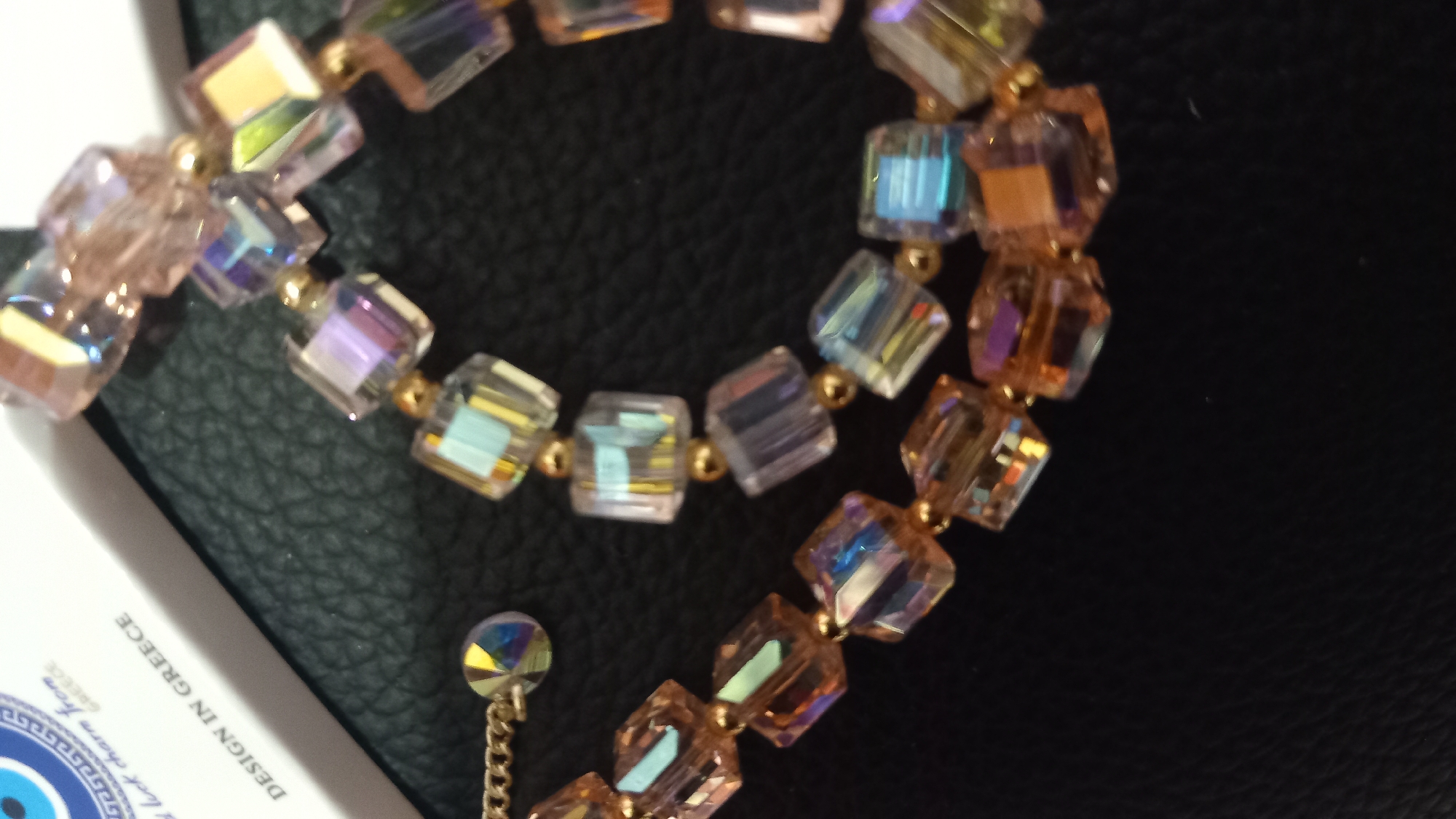 The image is a close-up, high-angle shot of a bracelet made of clear, faceted cubic beads with iridescent pink, blue, and yellow hues, separated by small gold-colored spherical beads. The bracelet is laid out on a textured black surface, possibly leather. In the upper left corner, a portion of a white rectangular card is visible, with text that reads "DESIGN IN GREECE" and a portion of a Greek evil eye charm with a blue and white circular pattern. The lighting creates reflections and highlights on the beads, showcasing their multifaceted nature. No people or specific locations within the city of Kos, Greece, are depicted, only the product itself and its packaging.