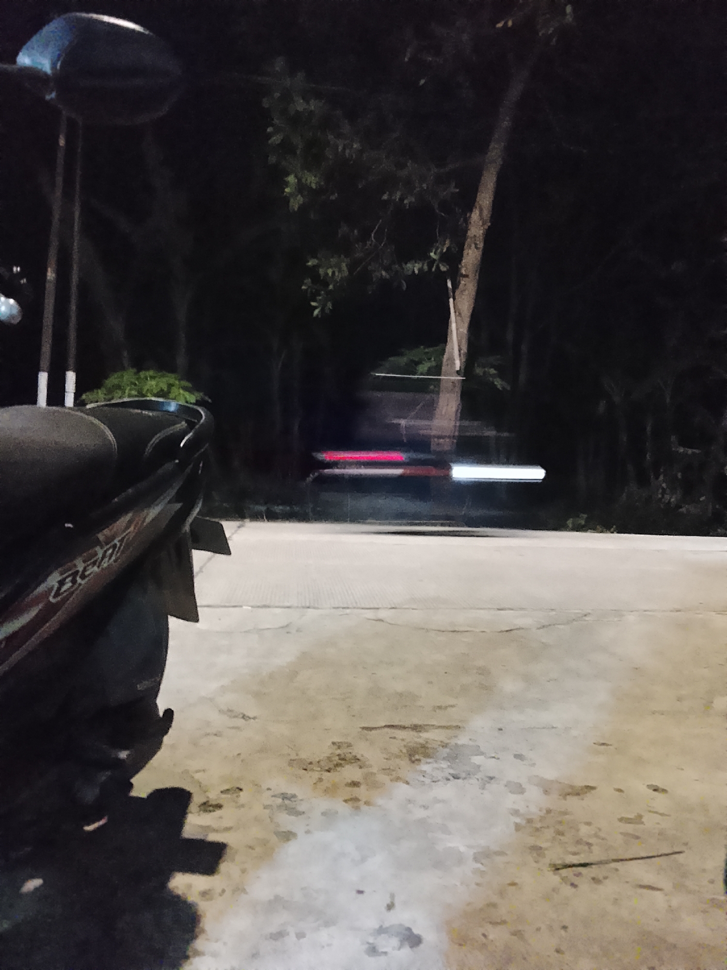 A motorcycle is parked on a concrete surface at night in Kalijaran, Indonesia. The motorcycle, partially visible from the rear, is dark in color with a red and white decal. In the background, a tree trunk is prominent, with dark foliage and trees surrounding it. A light streak, likely from a moving vehicle's headlights or taillights, is visible across the road. The concrete surface appears wet or stained in patches, and a shadow of the motorcycle is cast on the ground. The scene suggests a quiet, dark environment, possibly a roadside or a parking area.