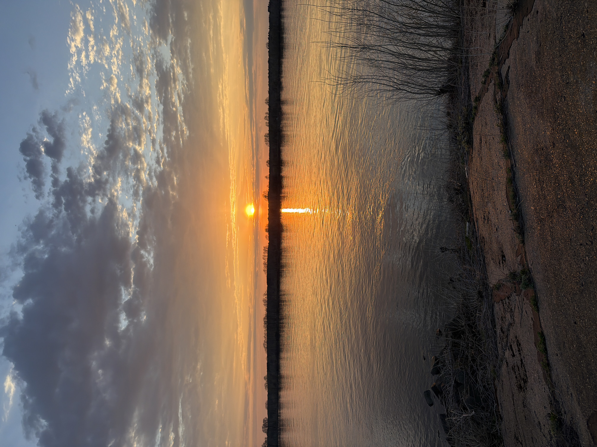 The image captures a sunset over the Kama River in Chaikovsky, Russia. The sun is centered on the horizon, with its reflection stretching across the water. The sky exhibits a gradient of colors, transitioning from blue with scattered clouds to orange and yellow near the horizon. Silhouetted trees line both sides of the river. In the foreground, on the right side of the frame, is a concrete embankment with sparse vegetation. The water surface is rippled, reflecting the sunset's colors.