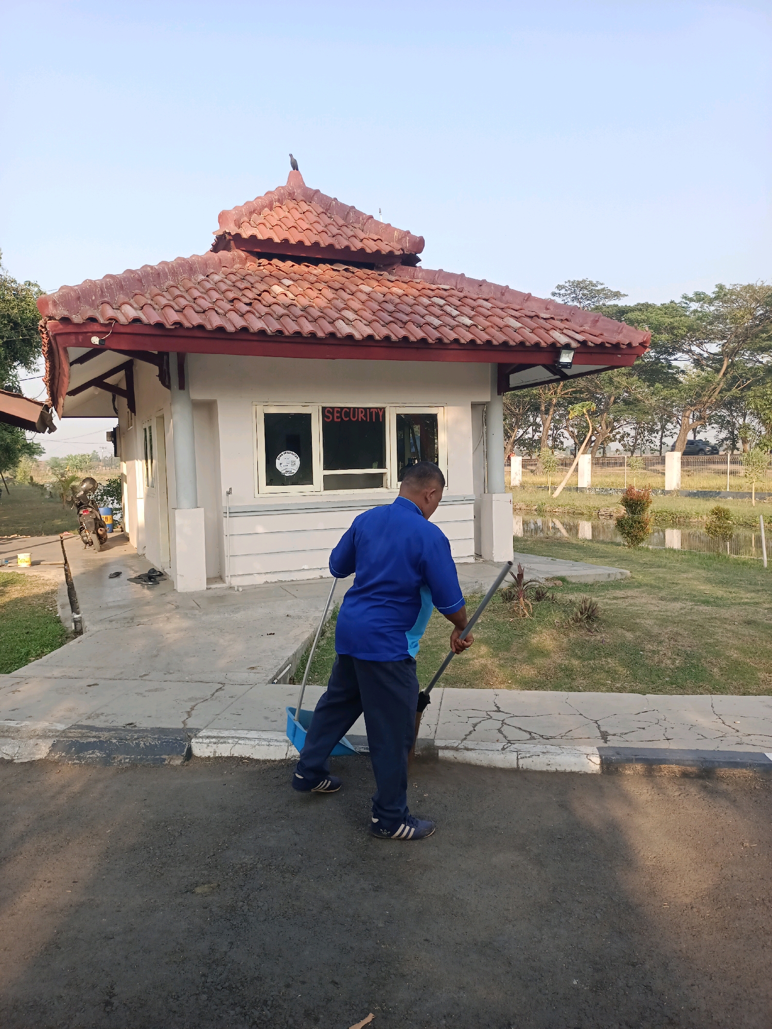 In Subang, Indonesia, a male security guard in a blue uniform is sweeping the paved area in front of a small security booth. The booth has a red tiled roof, a white exterior, and a window with the word "SECURITY" in red letters. The security guard is using a broom and a blue dustpan, collecting debris from the ground. The surrounding environment includes a grassy area, trees, and a small body of water. A motorcycle is parked to the left of the booth. The sky is clear and blue.