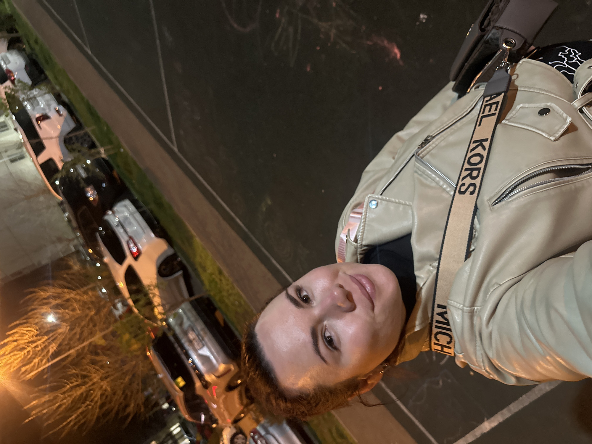 A woman stands on what appears to be the edge of a sidewalk or walkway in an urban environment. She is wearing a light-colored leather jacket and a bag with a shoulder strap labeled "MICHAEL KORS". Behind her is a row of parked cars along the curb, and the scene is dimly lit, suggesting it is nighttime. The location appears to be the city of Proletarskii, Russia.