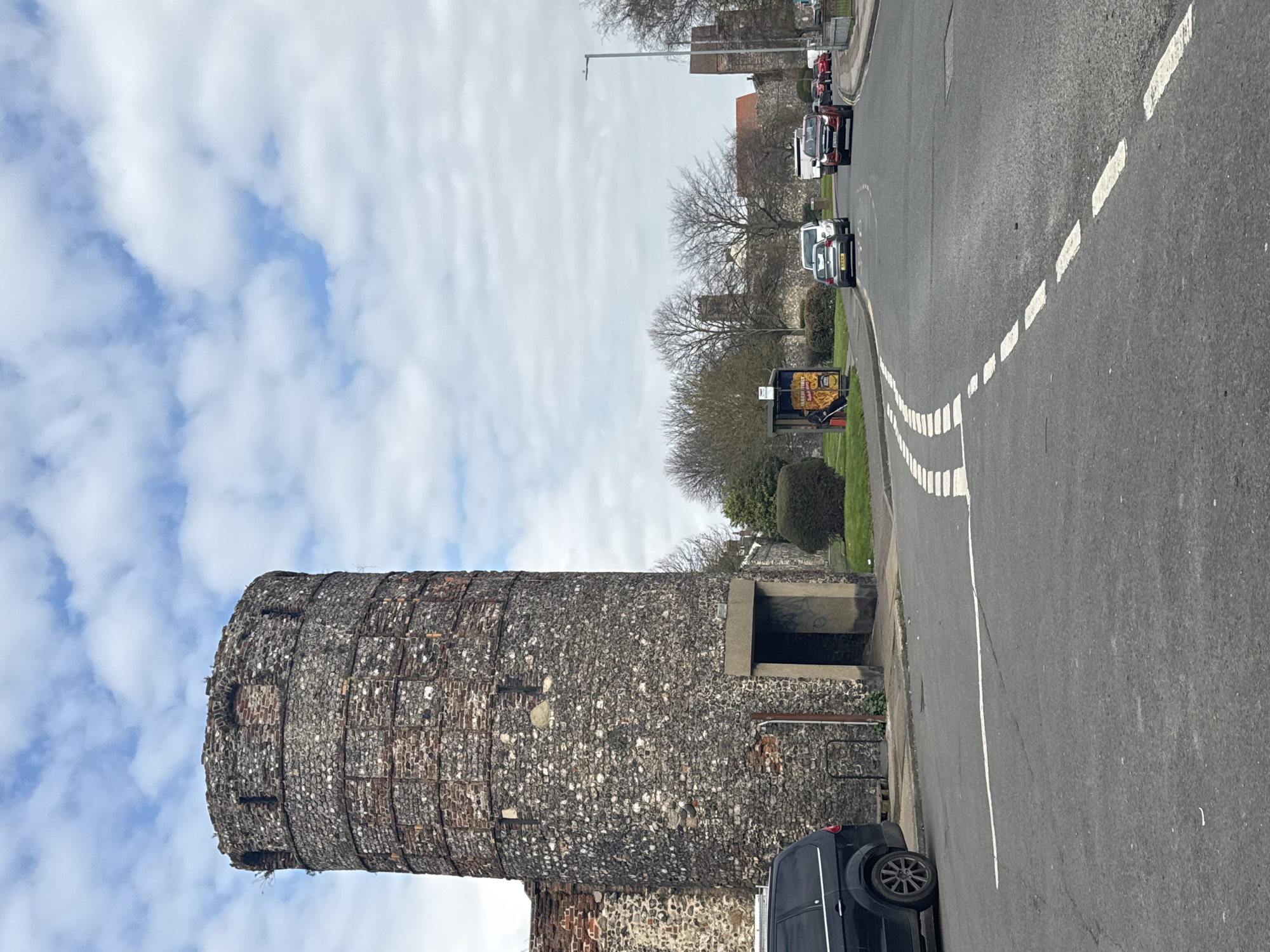 The image shows a stone tower, possibly part of a historical fortification, located near a road. The tower's exterior is constructed of stone and brick, with visible signs of age and repair. A dark-colored vehicle is partially visible in the foreground, parked along the roadside. The road has white dashed lines marking lanes or boundaries. A bus stop with an advertisement is present near the tower. The background shows a street with cars and buildings. The sky is partially cloudy. The image is taken in Gorleston-on-Sea, United Kingdom, where a preserved medieval flint tower is located on Gorleston High Street.