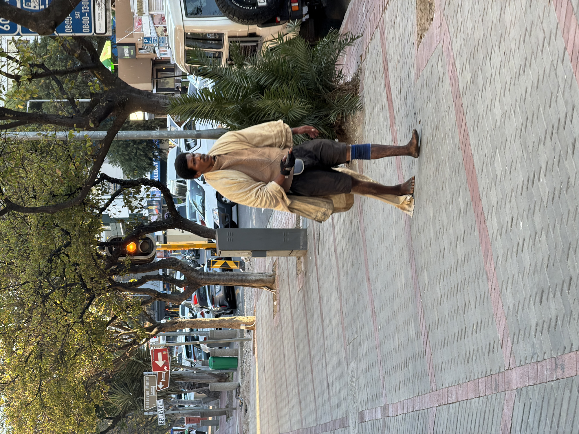 A barefoot African man is walking along a paved sidewalk, with his right foot slightly elevated, near a street filled with cars and trees in what appears to be Cape Town, South Africa. He wears dark-colored shorts, a tan shirt, and a loosely draped tan shawl or blanket. A blue bandage is visible on his left knee. He holds an object in his left hand, possibly a container or mobile phone. The sidewalk is made of gray brick, with red-brick horizontal stripes running along its length. Along the street, there are trees with yellowing leaves, traffic lights, and signs indicating "Bellevue Manor" and "Bellevue". Parked vehicles are visible. The overall environment suggests an urban setting, likely in a business or residential district of Cape Town.