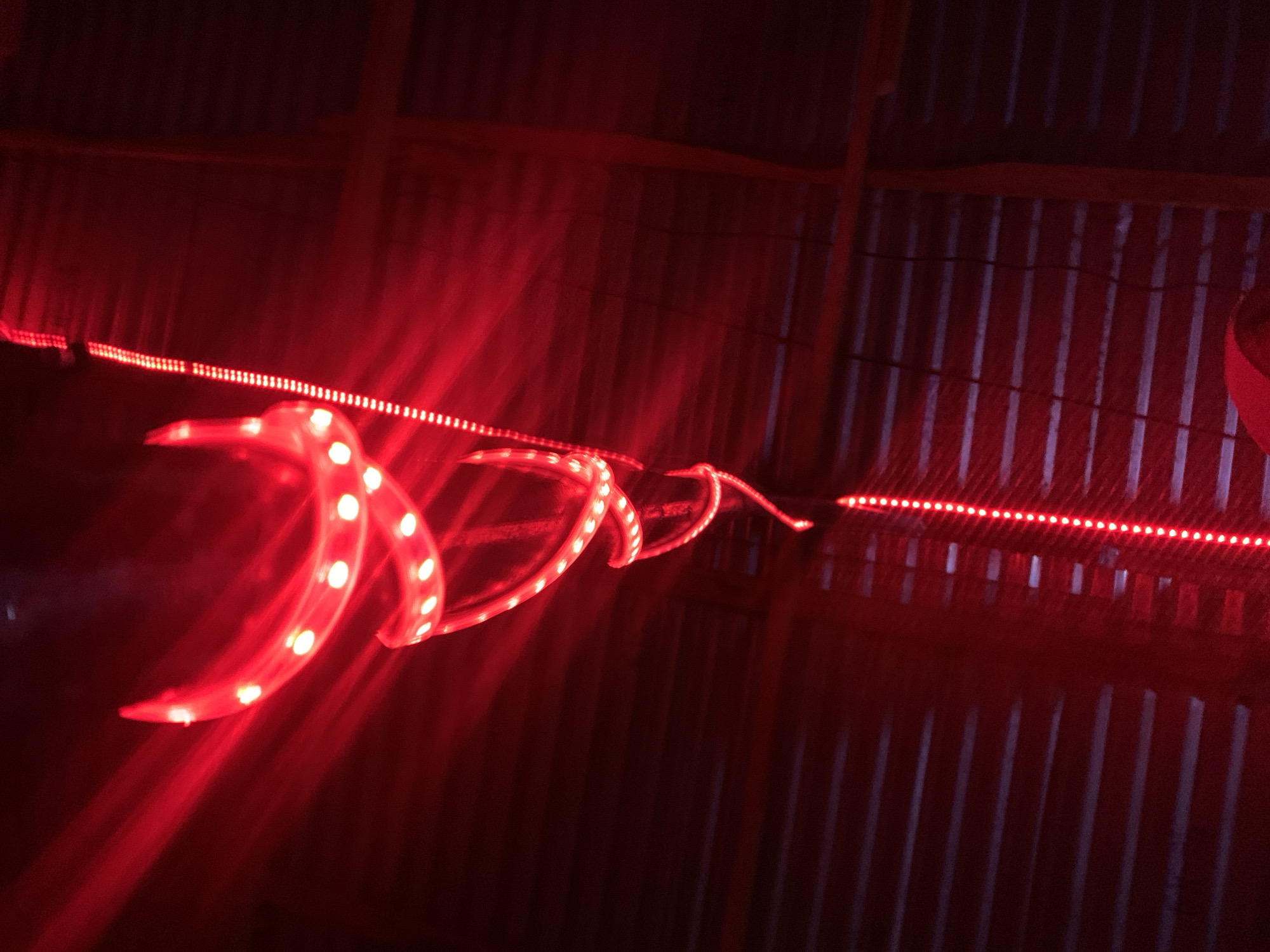A long exposure photograph shows a red LED light strip curled in a spiral. The strip is attached to the corrugated metal ceiling of a dark interior space. A straight red LED strip is attached along the metal. The image was taken in a dimly lit environment with a red tint. The location is assumed to be in Kabong, Nigeria.