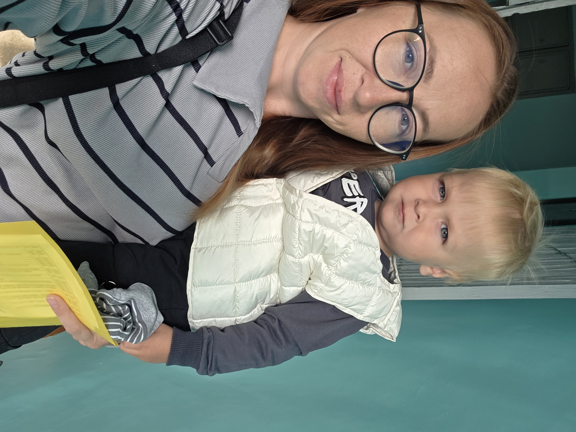 The image shows a woman holding a young child. The woman has long auburn hair and is wearing glasses and a gray and black striped shirt. The child has blonde hair and is wearing a white puffer vest over a dark long-sleeved shirt and black pants. The child is holding a pair of striped socks in their hands. The background is a teal-colored wall. There are no clear indications of the location being in the city of Chaikovsky, Russia.