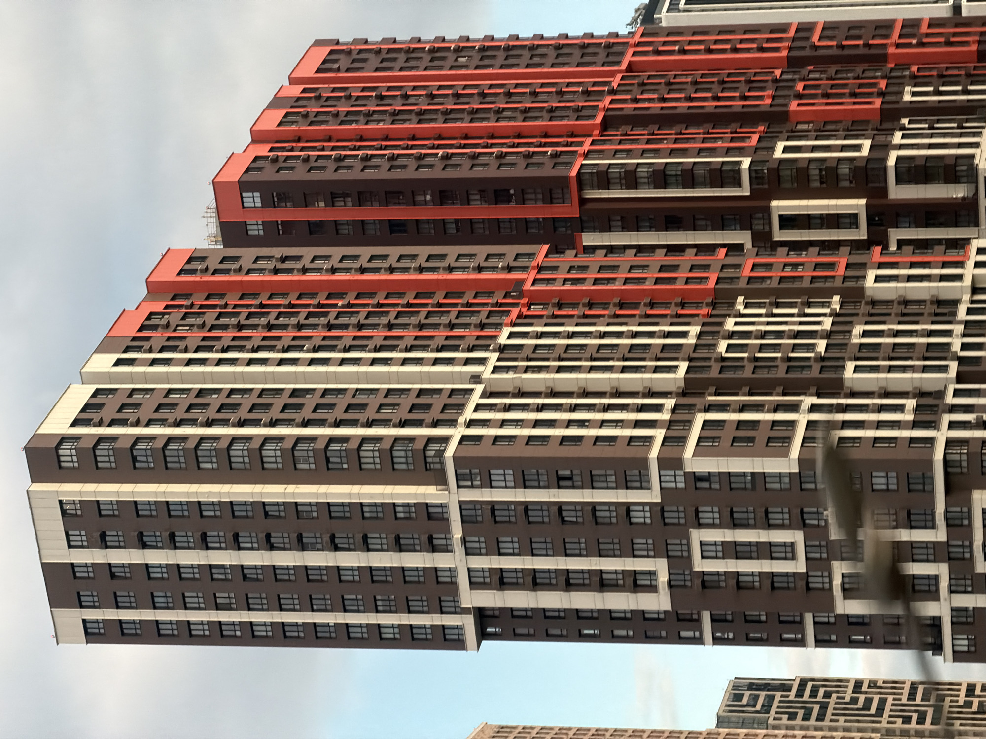 The original image, taken in Moscow, Russia, displays a towering, modern building with a distinctive and complex facade against a light, overcast sky. The primary subject is this multi-story building, which appears to be a residential or office complex due to its numerous, uniformly spaced windows.
The building's architecture is striking, characterized by a stepped or terraced design that incorporates bold geometric patterns and contrasting colors. The main sections of the facade are a dark brown, punctuated by numerous rectangular windows with dark frames. These dark sections are intricately interwoven with vibrant orange-red horizontal bands and off-white or light grey vertical and horizontal elements, creating a dynamic visual rhythm. Some sections of the building appear to cantilever or jut out, adding to its three-dimensional complexity. Small air conditioning units are visible beneath some windows, indicating occupied units.
In the lower part of the original image, partially obscured, another building can be seen with a different, intricate geometric pattern on its facade, resembling a labyrinth or maze.
The setting is an urban environment, with the building dominating the frame, suggesting a dense or developing city area. The time of day is daytime, evidenced by the clear, albeit muted, light from the sky, which is a pale blue-grey, indicating either overcast conditions or late afternoon. There are no people visible, and no specific activities or events are taking place, as the image focuses solely on the architectural structure. No visible text is present on the buildings. The overall impression is one of modern, large-scale urban development.