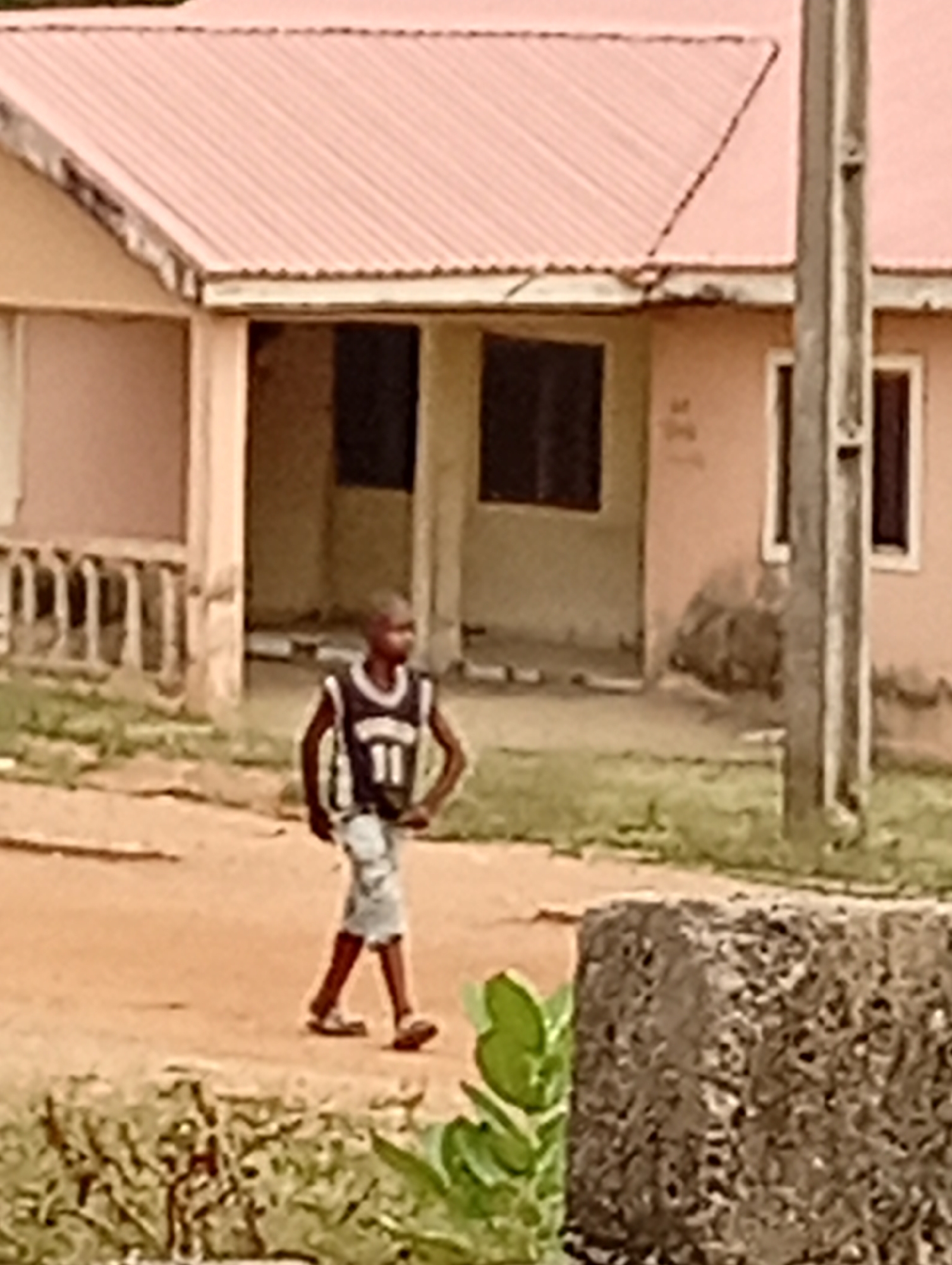 The image captures an outdoor scene in what appears to be a residential area, likely in Adankolo, Nigeria. The central subject is a young boy or man, positioned slightly left of center, walking or standing on a reddish-brown dirt road. He is wearing a dark sleeveless jersey with the number "11" clearly visible in white on the front, light-colored shorts, and flip-flops or sandals. His hands are placed on his hips, and his gaze is directed towards the right side of the frame.
In the background, two single-story buildings with light peach or pinkish-hued walls and reddish-pink corrugated metal roofs are prominent. The building on the left features a decorative white or light-colored balustrade or railing in front. Both structures have dark, rectangular windows. There are visible signs of wear and dirt on the lower sections of the building walls.
To the right of the frame, a tall, sturdy utility pole, possibly made of wood or concrete, stands upright. The ground is largely unpaved, with scattered patches of green grass and other low-lying vegetation. In the bottom right corner, a large, dark, textured concrete block is partially visible, with more green foliage in front of it. The lighting suggests it is daytime, possibly with an overcast sky, as there are no harsh shadows. The overall scene conveys a sense of everyday life in a developing community. The only distinct text visible is the number "11" on the subject's jersey.