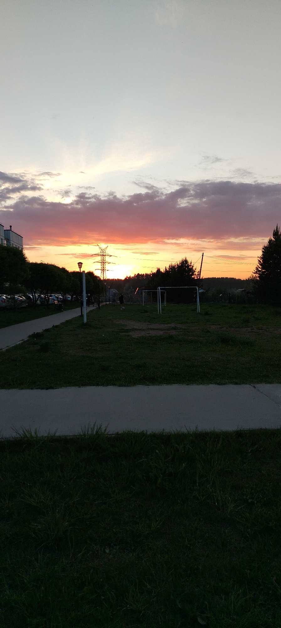 The image presents a landscape during sunset. The sky displays a range of colors from orange and pink near the horizon to lighter hues above. Clouds are visible near the horizon.
An electrical transmission tower stands silhouetted against the sunset. A soccer field is partially visible in the middle ground, with the soccer goal. There is a grassy area in front of the field and a paved walkway in the foreground.
People are seen near the soccer field. A multi-story building is on the left. Trees and other vegetation populate the middle distance.
This image was taken in the city of Chaikovsky, Russia.