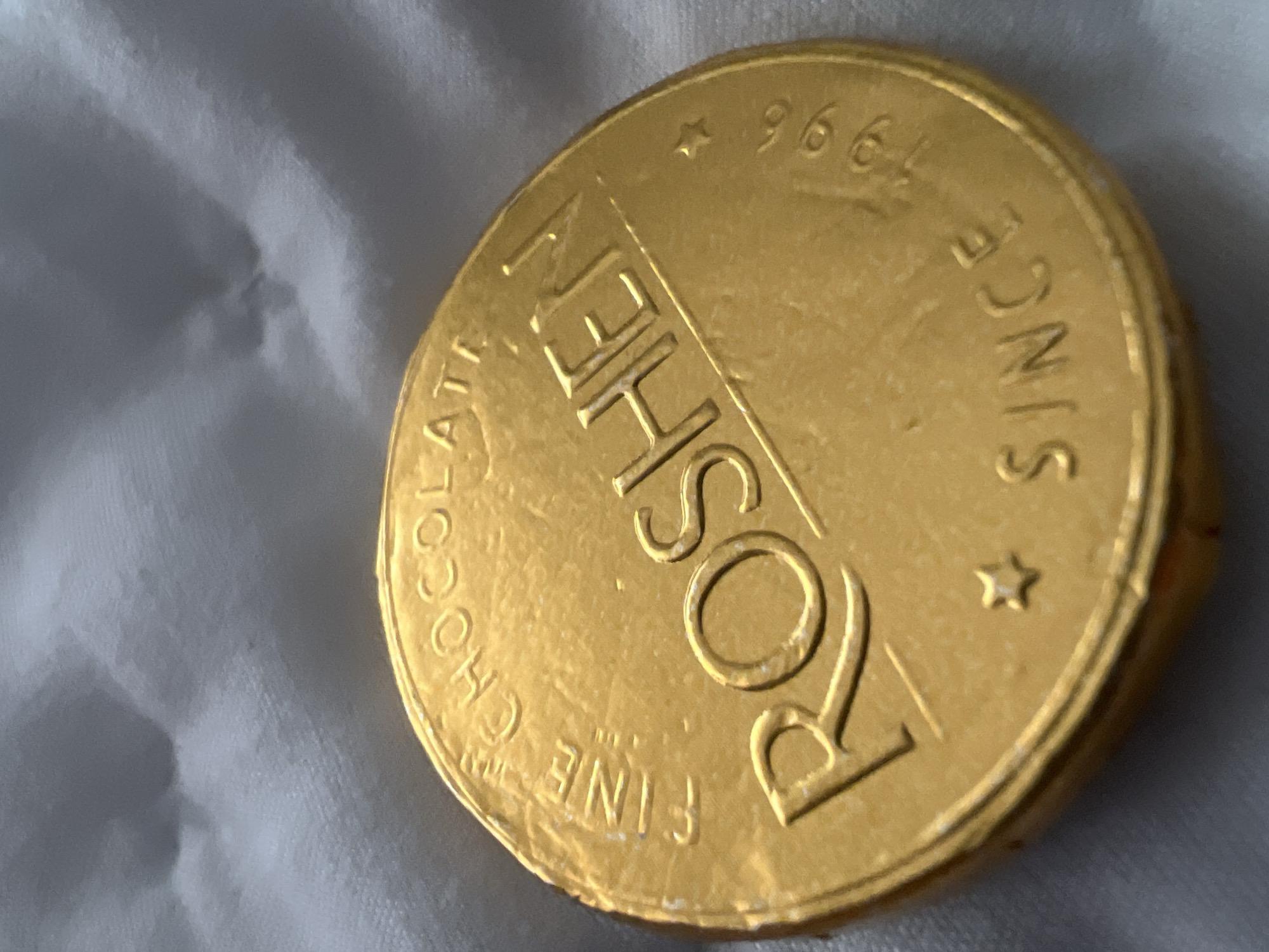A gold foil-wrapped chocolate coin with "ROSHEN FINE CHOCOLATE" and "SINCE 1996" inscribed on it rests on a white, textured surface. The object is not readily identifiable as being located in Budva, Montenegro based on the provided image.