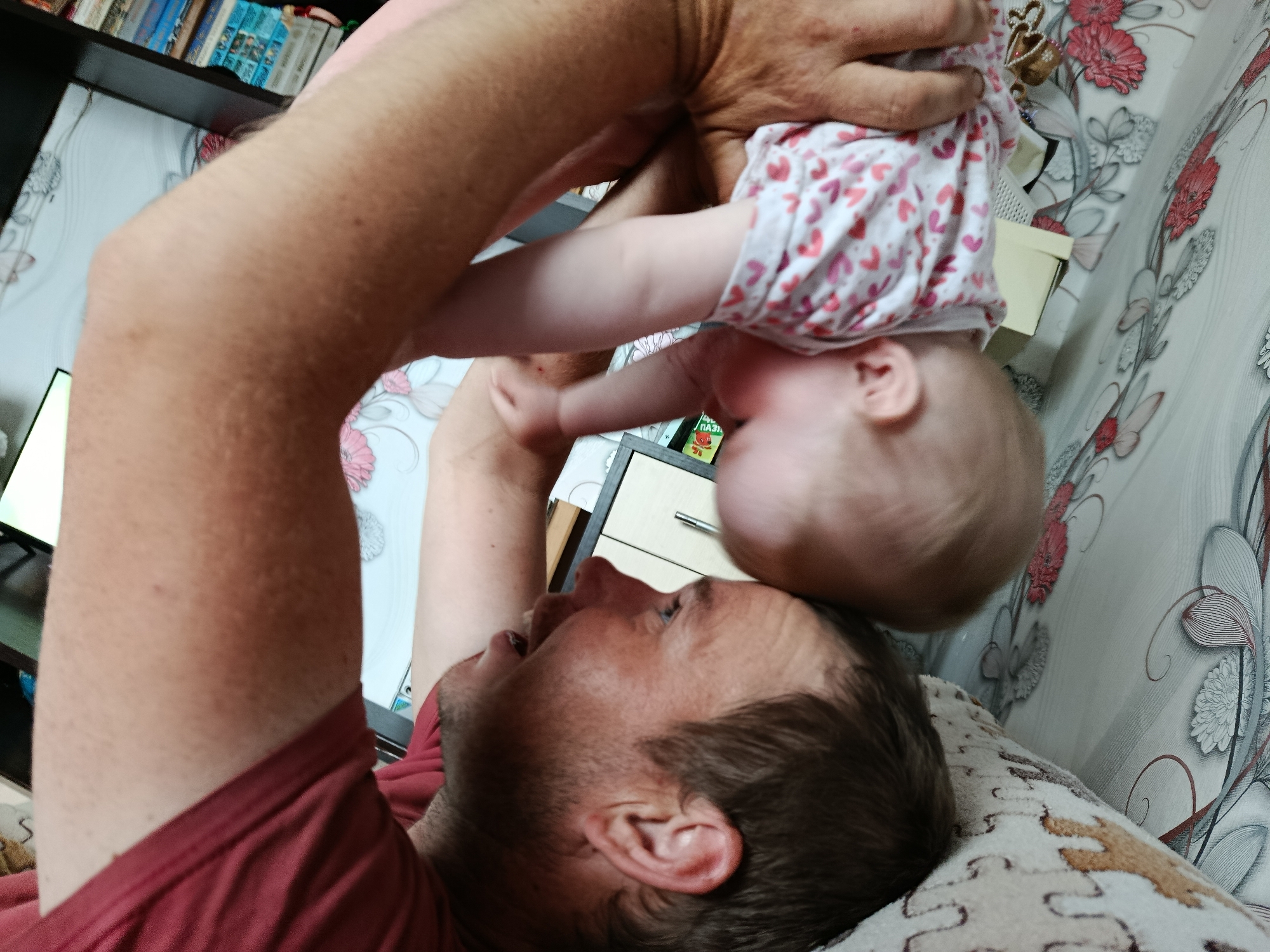A man lies on his back, lifting a baby in the air. The baby wears a white shirt with pink hearts. The man is light-skinned and wears a red t-shirt. They are indoors, lying on a patterned blanket, with a floral patterned wallpaper in the background. A shelf full of books is visible to the left. This image was taken in Фоки, Russia.
