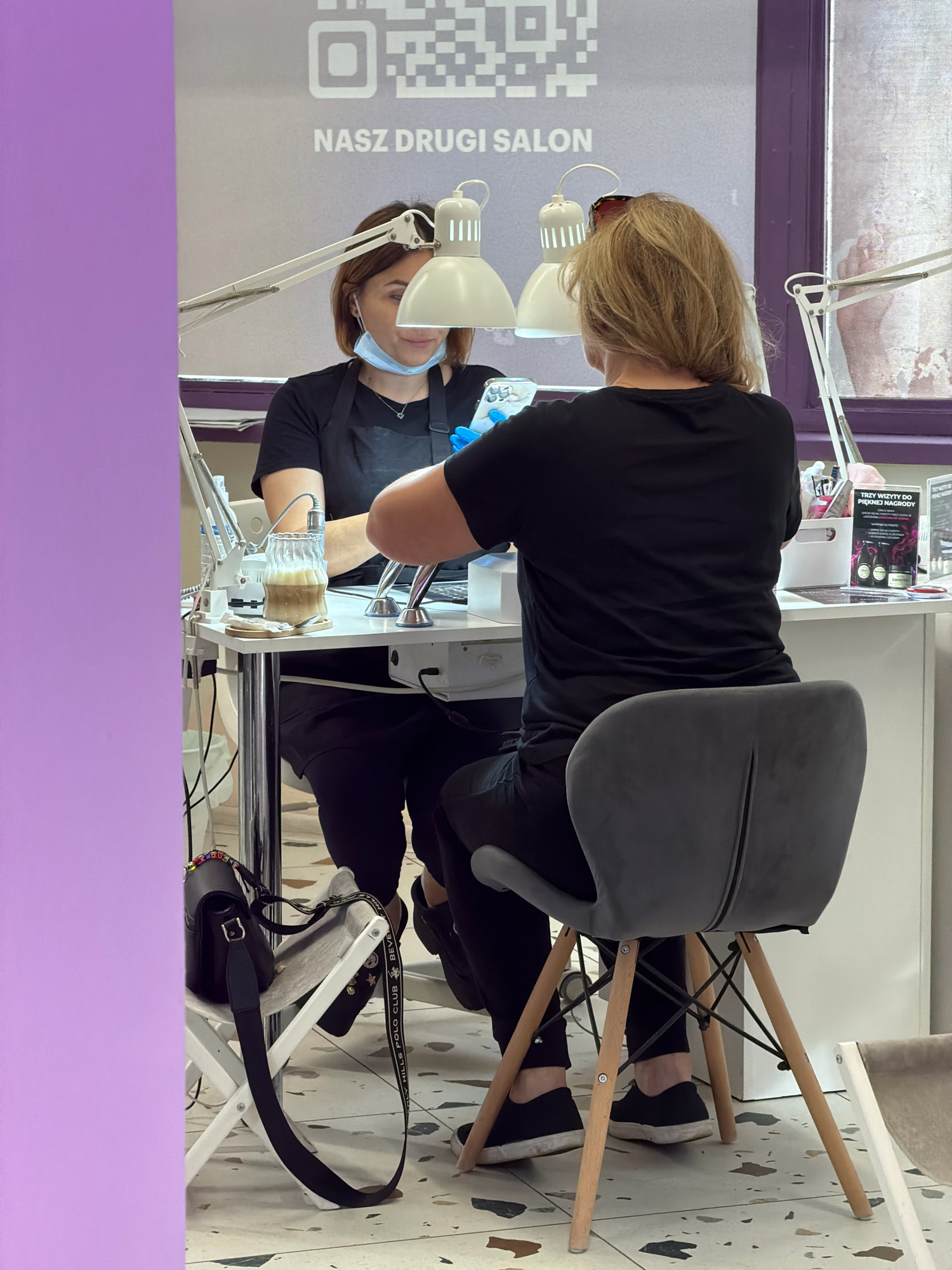 The image shows two women in a nail salon, likely located in Wrocław, Poland, given the Polish text "NASZ DRUGI SALON" (Our Second Salon) visible on the wall behind them, alongside a QR code. The woman on the left, wearing a black apron and a light blue face mask hanging from her ear, is attending to the nails of the woman on the right. Both women are seated at a white table. The woman on the left has a cup of coffee and various manicure tools on the table in front of her. The woman on the right is wearing a black t-shirt and black pants, and is seated in a modern grey chair with wooden legs. In the background, there are several lamps, a white filing cabinet, and promotional materials, including one that advertises "TRZY WIZYTY DO PIĘKNEJ NAGRODY" (Three Visits for a Beautiful Reward). To the left of the table, a folded white chair holds a black purse with a colorful beaded strap and a black strap with white lettering. The floor has a speckled terrazzo pattern.