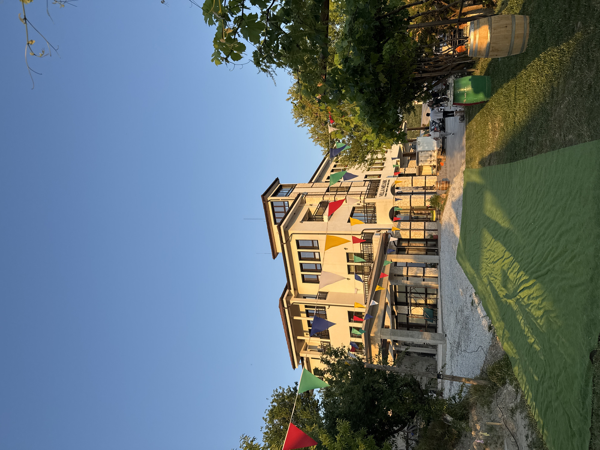 The image displays a multi-story building, identified by a sign on its facade as "VILLA MELNIK FAMILY WINERY AND VINEYARDS," located in Harsovo, Bulgaria. The building features a cream-colored exterior, brown-tiled roofs, multiple dark-framed windows, and balconies with dark railings. Colorful triangular pennant flags, including red, green, blue, yellow, and white, are strung across the building's front and among the surrounding trees. In the foreground, a strip of bright green artificial turf or manicured grass runs diagonally from the lower left to the mid-right, displaying prominent long shadows. To its left, the ground consists of light-colored gravel or crushed stone. To the right, natural green grass with visible shadows extends towards the building. Lush green trees and foliage frame the scene on both sides, with dense grapevines visible on the right. Two wooden barrels, one upright and one on its side, are positioned on the grassy area on the right, along with a green metal container. Several people are visible near the building's entrance/driveway on the right; one individual in light clothing walks towards the right, while others are gathered near a white utility vehicle and what appears to be an outdoor serving station with tables. The scene is illuminated by low, warm sunlight, casting long shadows, indicative of late afternoon or early evening. The sky above is clear and light blue.