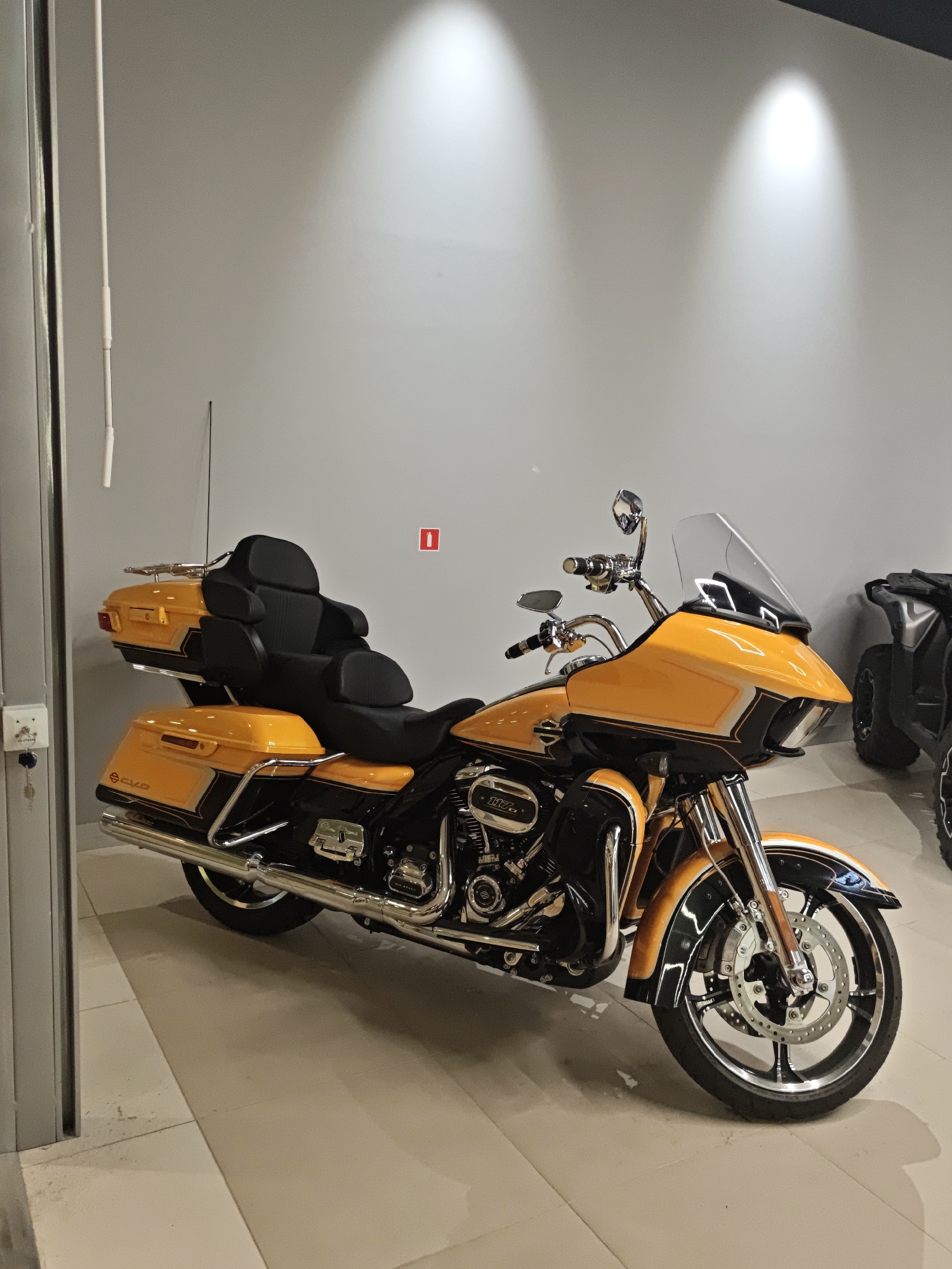 This is a full-frame, eye-level, medium shot of a bright orange and black motorcycle parked indoors. The motorcycle is the primary subject and is angled slightly towards the viewer's right. It features a large windshield, a comfortable-looking dual seat with a prominent backrest, and two saddlebags. The motorcycle has a chrome exhaust system and intricate detailing on its engine.
The setting appears to be a showroom or garage, indicated by the smooth, light-colored tiled floor and the neutral gray walls. Two spotlights illuminate the upper portion of the wall, casting distinct pools of light. On the left side of the frame, a gray metal column and a white electrical conduit are visible. A small red square sign with a white icon is on the wall behind the motorcycle's seat.
The motorcycle is stationary, and no people are present. The lighting suggests it is daytime, likely within business hours given the showroom-like environment. The overall mood is clean and professional, highlighting the vehicle. Visible text includes "117" on the engine and a logo on the saddlebag that appears to be "CVO". A small white box with a key attached is mounted on the gray column on the left. Another vehicle, possibly an ATV, is partially visible in the background on the right.