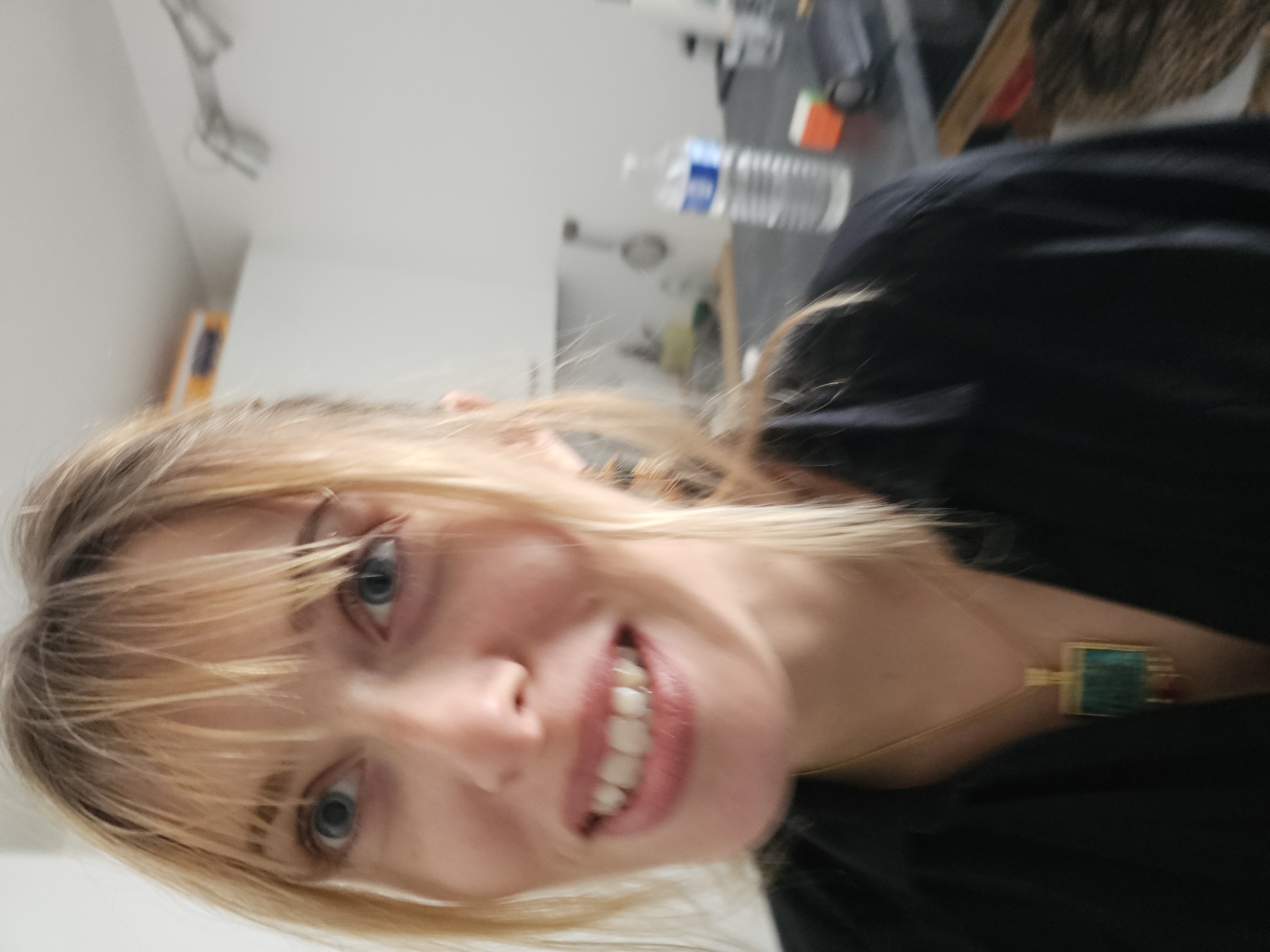 A selfie of a blonde woman with blue eyes, wearing a black top and a gold necklace with a rectangular green and red pendant, is centered in the frame. The background is blurred and appears to be an interior space. A water bottle and other objects are visible on a countertop in the background. Based on this information, there is not sufficient detail to place the subject in Pantin, France.