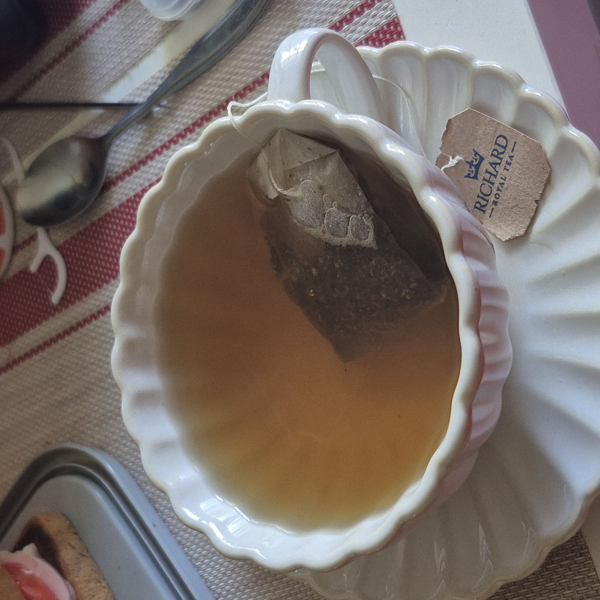 The image shows a close-up view of a cup and saucer filled with tea. A tea bag is immersed in the tea, and a small tag with the text "RICHARD - ROYAL TEA -" is attached to the side of the cup. In the background, a spoon rests on a red and white striped tablecloth. To the left of the cup, a portion of a sandwich or pastry is visible on a grey plate. The scene appears to be indoors, possibly in a home or cafe setting, and there is no indication of the city of Chaikovsky, Russia.