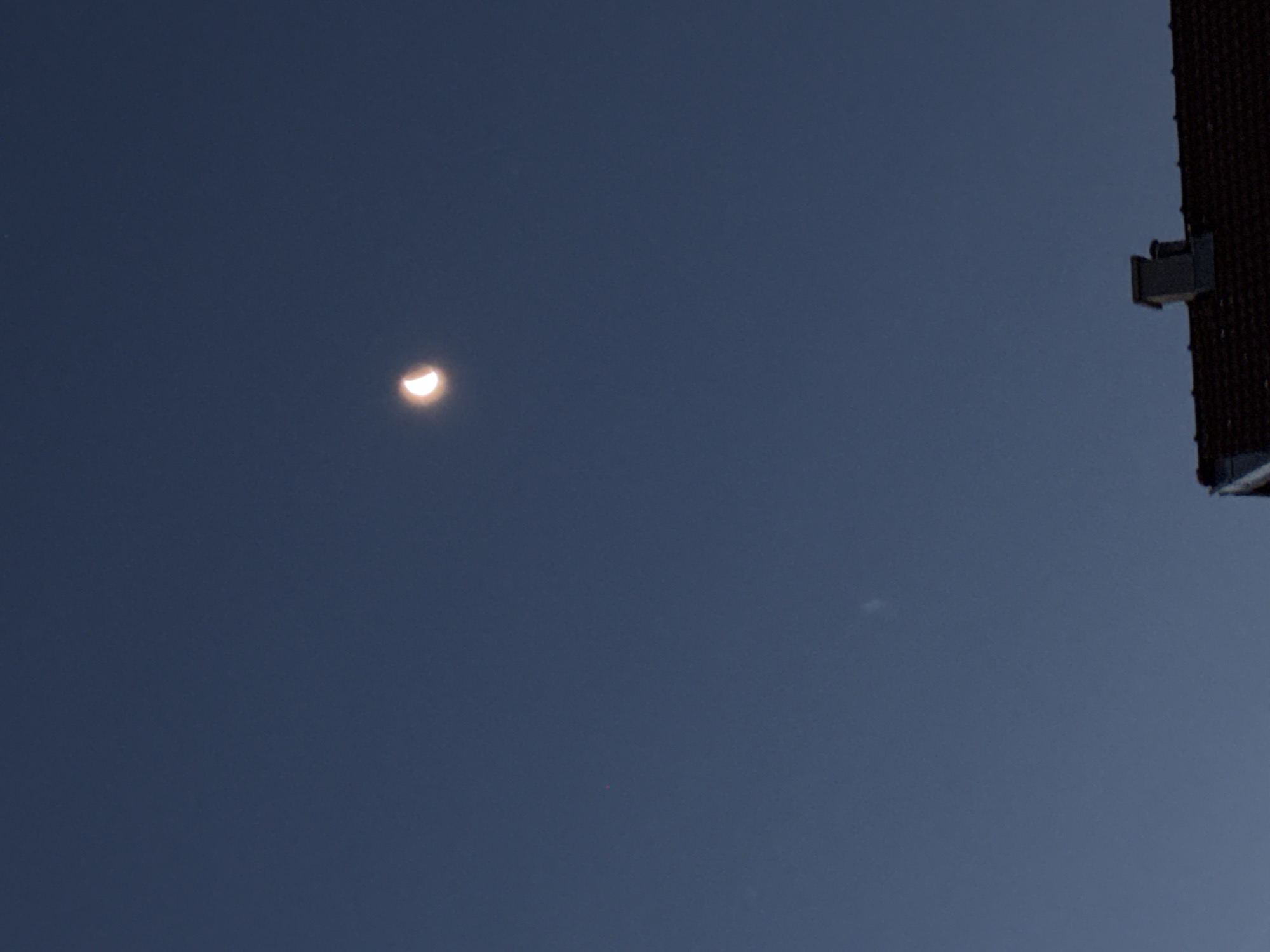 The image is a night or twilight sky with a crescent moon and a faint smudge of light, possibly a distant cloud or atmospheric phenomenon, located in the upper half of the frame. In the lower portion of the image, the roofline of a building is visible, with a chimney. The setting is outdoors, suggesting an urban or suburban environment given the presence of the building. The lighting indicates it is not broad daylight.