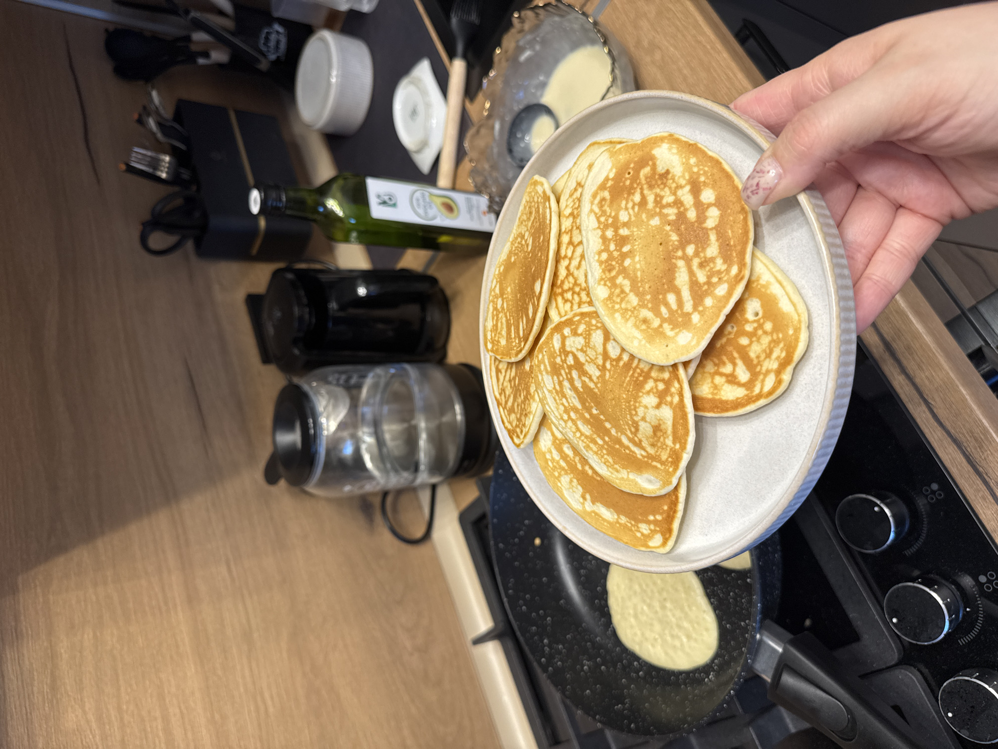 A close-up, high-angle shot depicts a kitchen setting, presumably indoors. A light-skinned hand holds a gray plate piled with golden-brown pancakes. Below the plate, a frying pan sits on a black stovetop, containing a single pancake in the process of cooking. Various objects are scattered on the wooden countertop to the left of the stovetop, including a glass bottle of oil, a black electric kettle, and a container of pancake batter. The overall impression is one of breakfast preparation. The location in Yakhroma, Russia, is not discernible from the visual information provided.