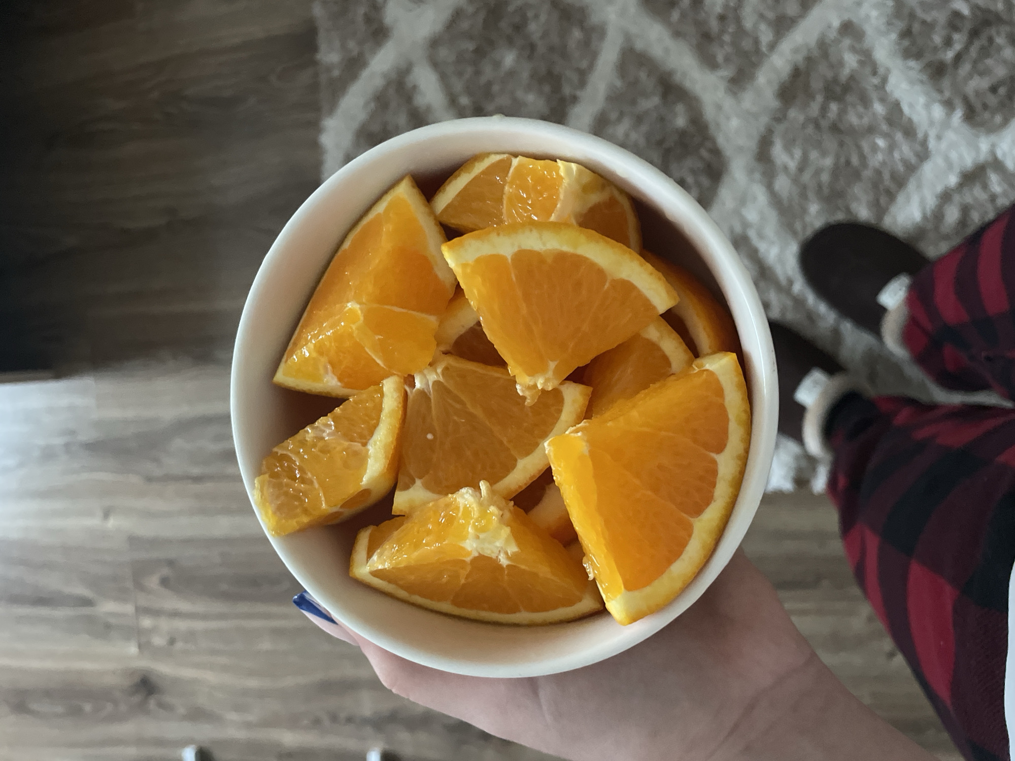 A person is holding a white bowl filled with numerous bright orange slices. The bowl, which appears to be ceramic, is held in the center of the frame by a hand with blue nail polish on the thumb. The orange segments are cut into wedges, showing their juicy pulp and some white pith.
The individual is wearing red and black plaid pajama bottoms or lounge pants, and dark-colored, possibly brown or black, fuzzy slippers are visible on their feet. The setting is indoors, featuring a light brown wooden floor on the left side of the frame and a light-colored, possibly off-white or gray, patterned rug on the right. The lighting suggests it is daytime. The overall impression is one of a relaxed, casual moment at home, enjoying a fresh fruit snack. No text is visible in the image. The scene is set in Zlatograd, Bulgaria.