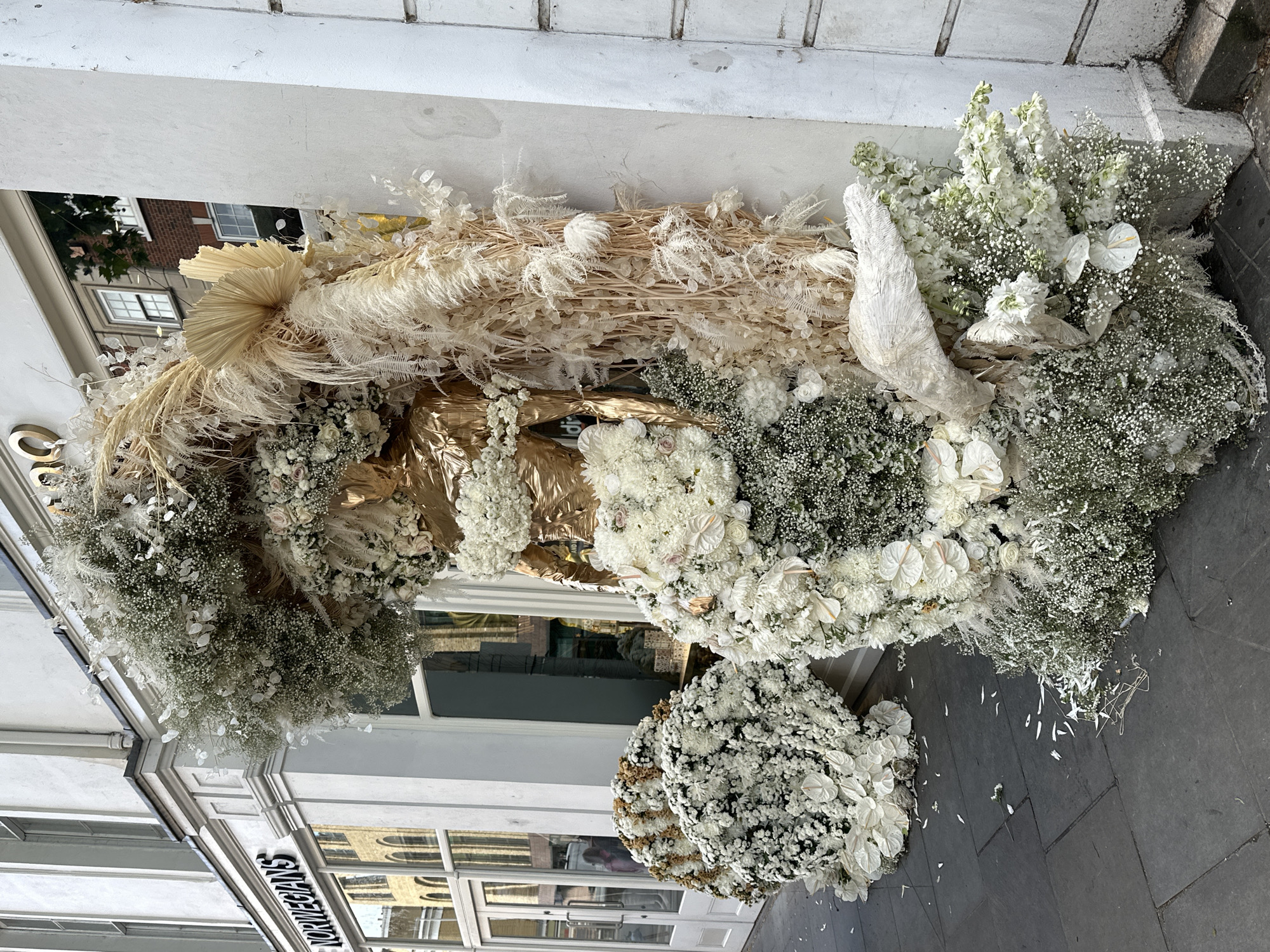 The image shows a lavish floral arrangement outside a building, likely in London, United Kingdom. The display is a mix of white and cream-colored flowers, including baby's breath, roses, and what appears to be pampas grass. The arrangement is partially metallic gold, adding a touch of elegance and opulence. It is installed against a white wall, presumably the exterior of a store or building, and extends onto the sidewalk. The sidewalk is grey and littered with fallen petals and small flower pieces. The architecture visible in the background suggests a typical urban streetscape in London.