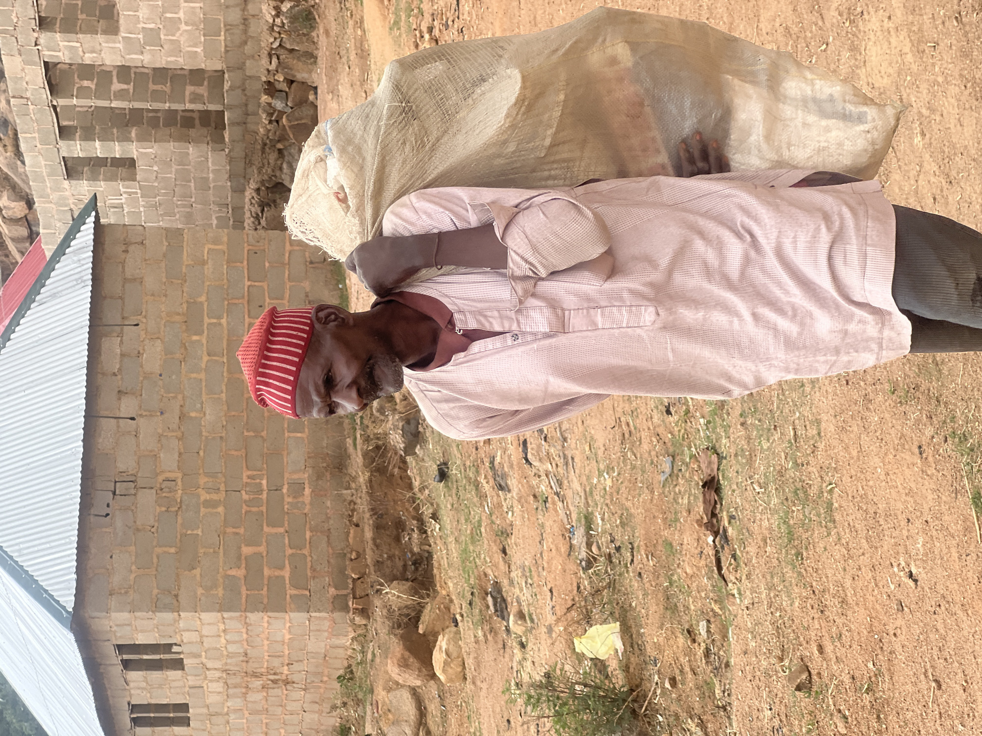 An elderly man with a red and white striped cap carries a large, beige-colored sack over his left shoulder. He wears a light pink, long-sleeved shirt and dark pants. The man is walking on a dirt and grass terrain. Behind him stands a building constructed of light grey cinder blocks, topped with a corrugated metal roof. The ground is littered with small rocks, sparse vegetation, and scattered pieces of trash. The location is outdoors. This image appears to be captured in a rural area in the city of Jos, Nigeria.