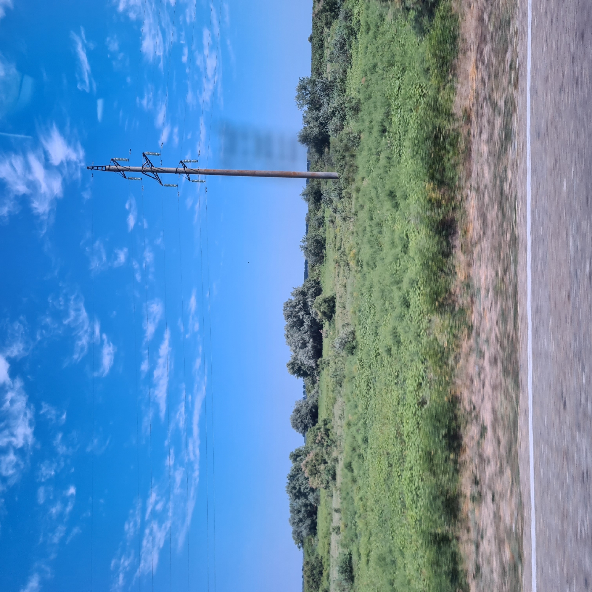 The image shows a rural landscape with a power pole in the foreground, a field of green grass and bushes, and a bright blue sky with some wispy clouds. The scene appears to be taken from a moving vehicle, as indicated by the blurred foreground. There are no people or animals visible in the image. The power pole is made of wood and has insulators and wires attached to it. The vegetation is dense and appears to be overgrown in some areas. The sky is clear with a few white clouds scattered across it. The overall impression is one of a quiet and serene natural setting. The location is identified as Zaton, Russia.
