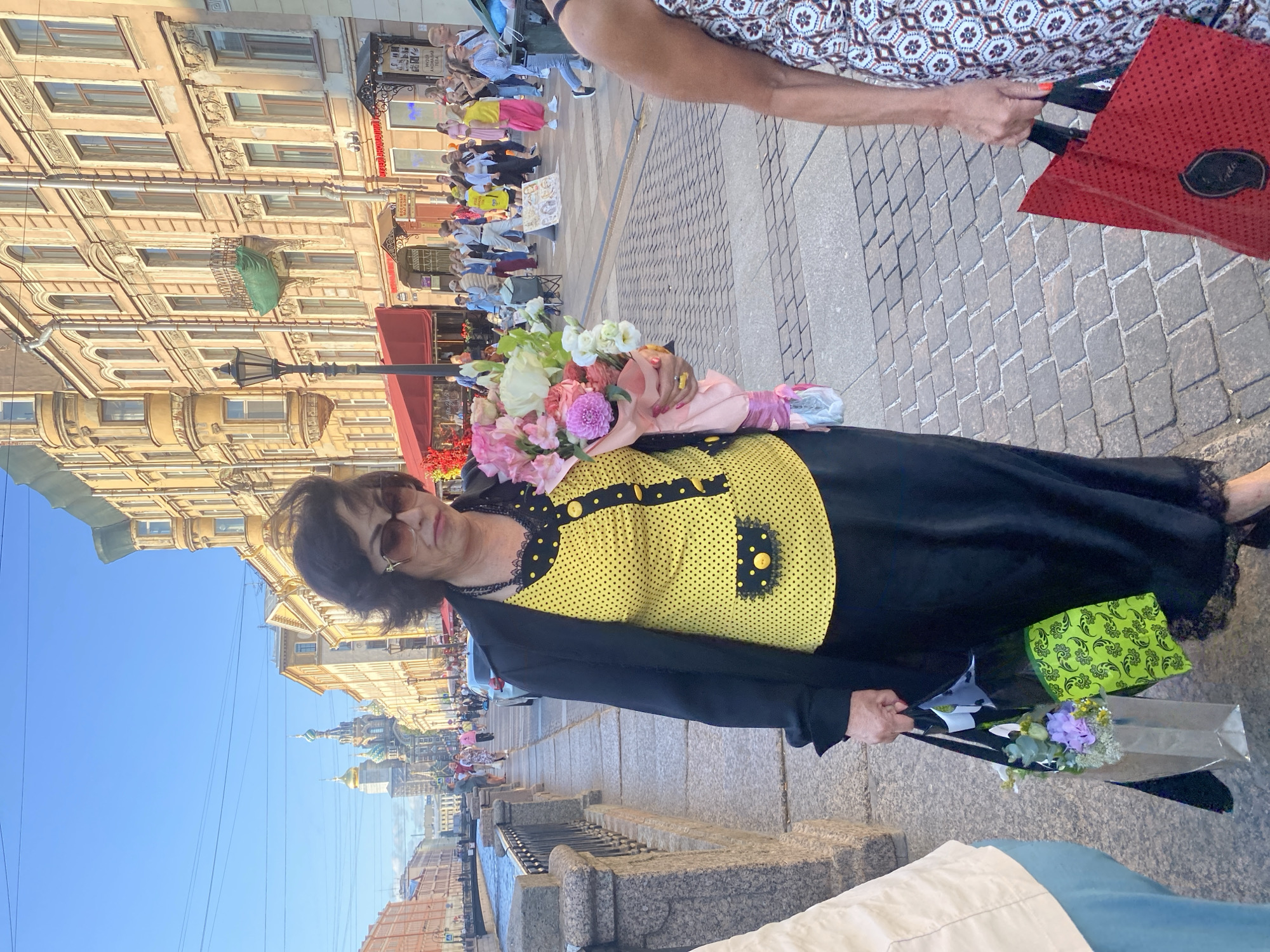 A woman with dark hair and sunglasses, wearing a yellow polka-dot blouse and black skirt, walks along a cobblestone sidewalk next to a canal in Saint Petersburg, Russia. She is holding a bouquet of flowers and two shopping bags. The background shows historic buildings and a crowd of people. In the distance, a church with golden domes is visible. The sun is shining, casting long shadows.