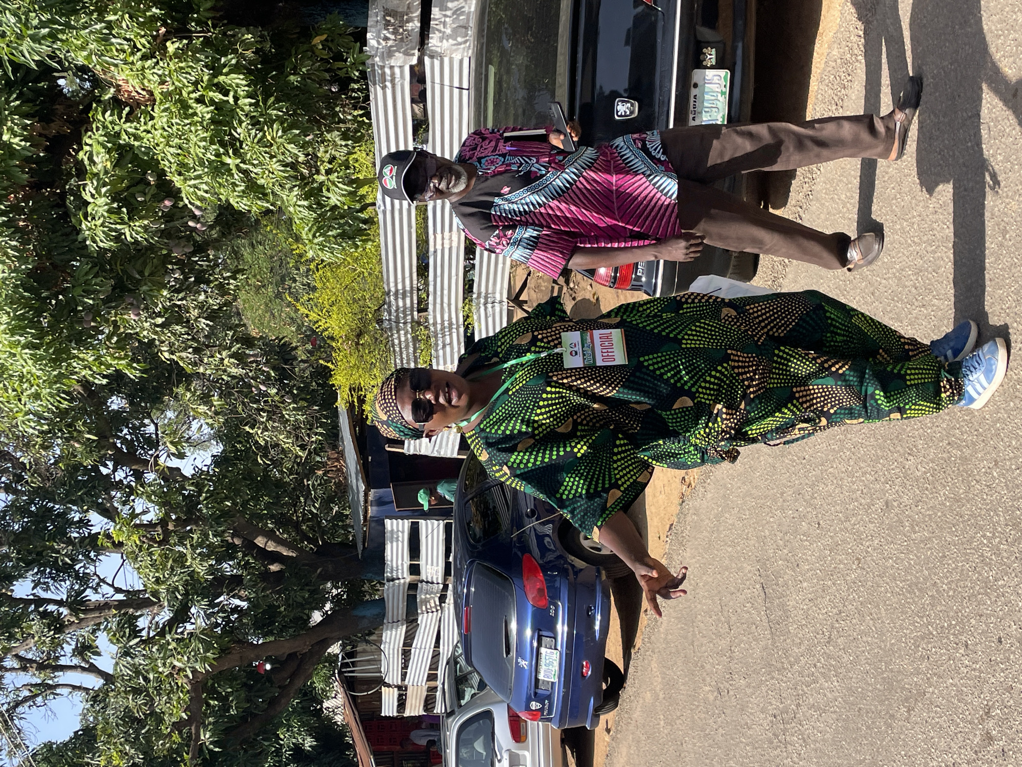 The image, captured in Jos, Nigeria, features two individuals standing outdoors on a sunny day. Strong shadows on the paved ground suggest it is midday or early afternoon.
In the foreground, a woman stands on the left, facing the viewer. She is wearing a vibrant green, black, and yellow patterned dress or traditional outfit, a matching headwrap, and sunglasses. An "OFFICIAL" lanyard with additional, smaller text that appears to say "Meta" is visible around her neck. Her left hand is slightly raised. Behind her is a dark blue Peugeot 206 hatchback with the Nigerian license plate "ABUJA RUM-965TU". A white car is partially visible behind the blue one.
To the right and slightly behind the woman, a man stands, also facing the viewer. He wears a striking pink, blue, and black patterned shirt, dark brown trousers, and a black baseball cap. He has a grey beard and is holding a black smartphone. Behind him is a black Peugeot sedan, partially showing its emblem and the Nigerian license plate "ABUJA BDG-994AAS".
The background is dominated by lush green trees, likely mango trees, with small fruits visible among the leaves. Simple structures made of corrugated metal sheets are also present. The scene depicts individuals possibly attending or participating in an event, indicated by the "OFFICIAL" tag, in a bright, outdoor setting.