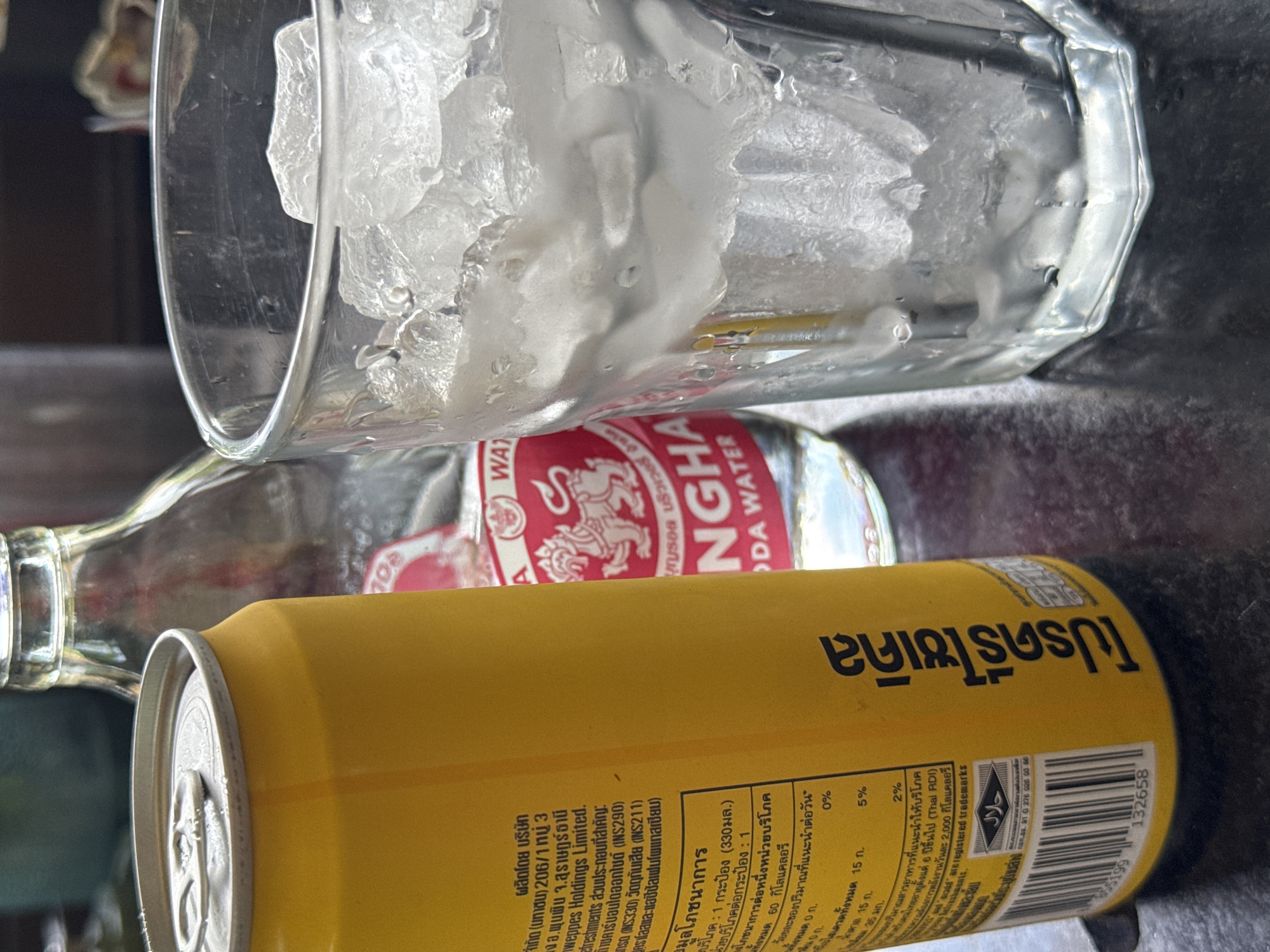 A close-up, slightly angled shot captures a yellow can of Singha soda water and a glass filled with ice and water. To the left, the yellow can is partially visible, showing text in Thai script. Behind the can, a clear glass bottle, also with a Singha label, is present. To the right, a clear glass is filled with a generous amount of crushed ice. The glass is sweating, indicating a cold beverage. The subjects are placed on a dark, reflective surface that appears to be a table, possibly outdoors or in a casual setting.