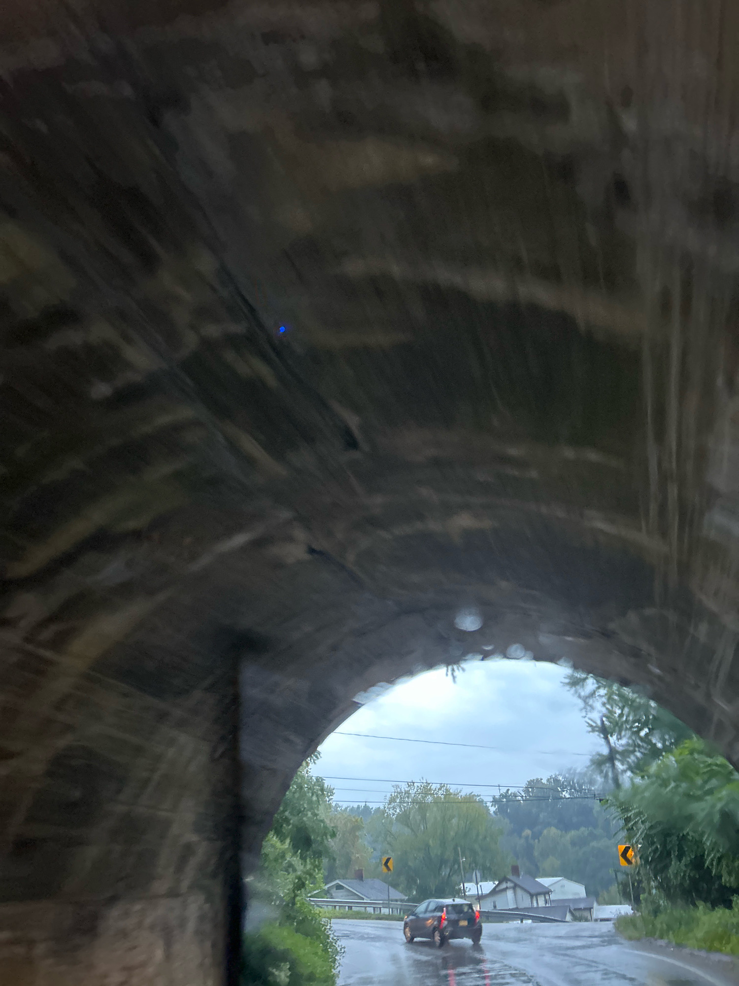 The image shows a view from inside a stone tunnel, looking outwards. The tunnel's rough-hewn stone ceiling dominates the upper portion of the frame, with visible striations and a small, bright blue light. The tunnel opens to a grey, overcast sky. Outside the tunnel, a wet asphalt road is visible, reflecting the dim light. A dark-colored SUV is driving away from the camera on the right side of the road, its brake lights illuminated. On either side of the road, there are yellow, curved arrow signs indicating a turn. The road is lined with houses and lush green trees, suggesting a semi-rural or suburban environment. The overall scene suggests a damp, possibly rainy day.