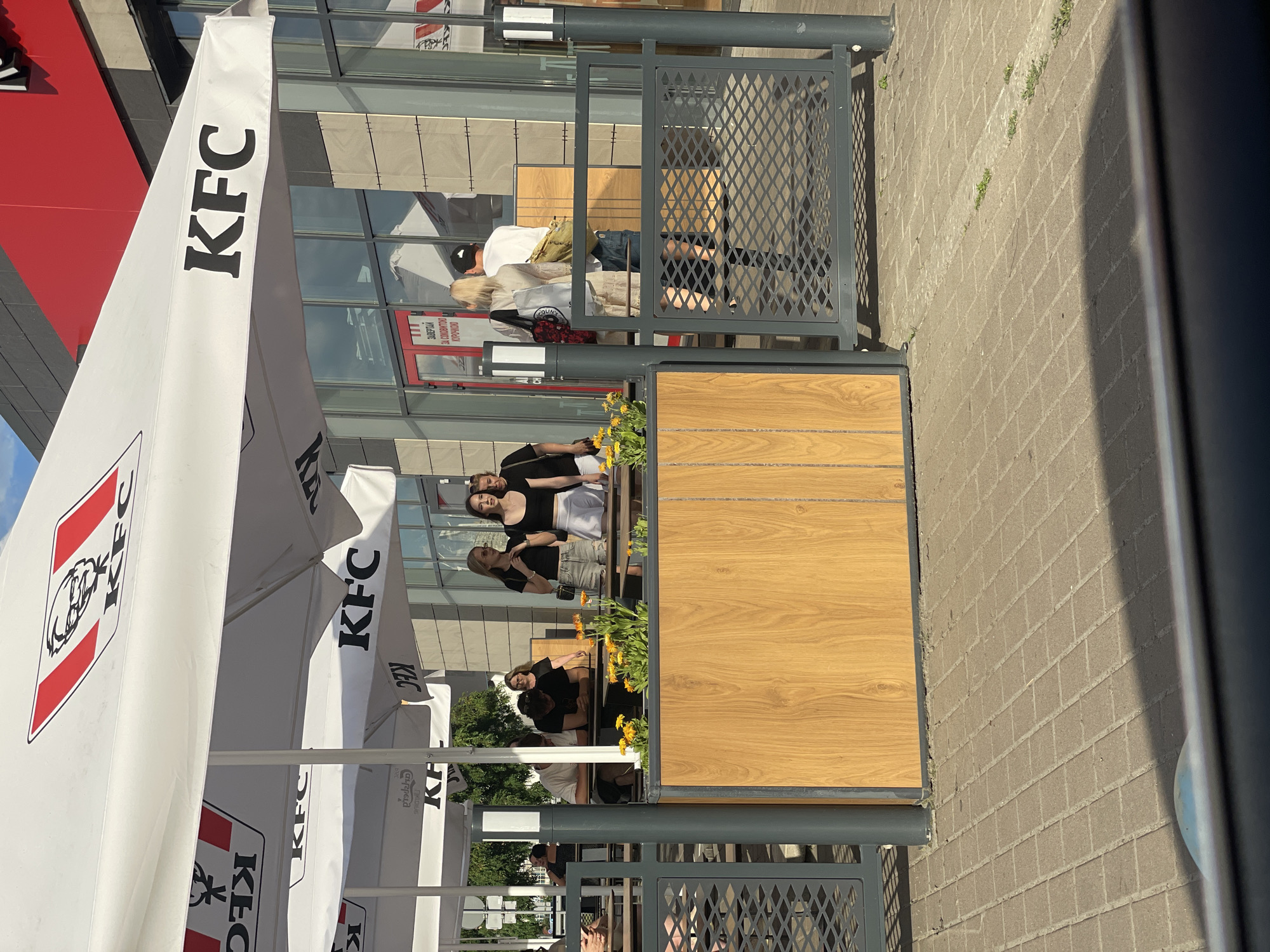 The image depicts an outdoor seating area of a KFC restaurant in Kyiv, Ukraine. The scene is bathed in sunlight, suggesting a bright, clear day. White KFC umbrellas are prominently displayed, providing shade over the tables. In the background, the KFC building with its distinctive red and white branding is visible, along with large glass windows reflecting the surroundings.
Several people are seated in the outdoor area, appearing relaxed and engaged in conversation or enjoying their meals. One group of women is standing and posing for a photo, with smiles suggesting a pleasant social interaction. Another person, a woman, is seated at a table, looking down at something in her hands. The overall mood seems casual and convivial.
The seating area features a modern design with wooden accents and metal railings. Planters with yellow flowers add a touch of color to the space. The ground is paved with brickwork. The time of day appears to be afternoon, judging by the angle of the sunlight and the shadows cast. Visible text includes "KFC" on the umbrellas and a sign on the building that includes Cyrillic script. The atmosphere is indicative of a typical day at a fast-food restaurant with outdoor seating.