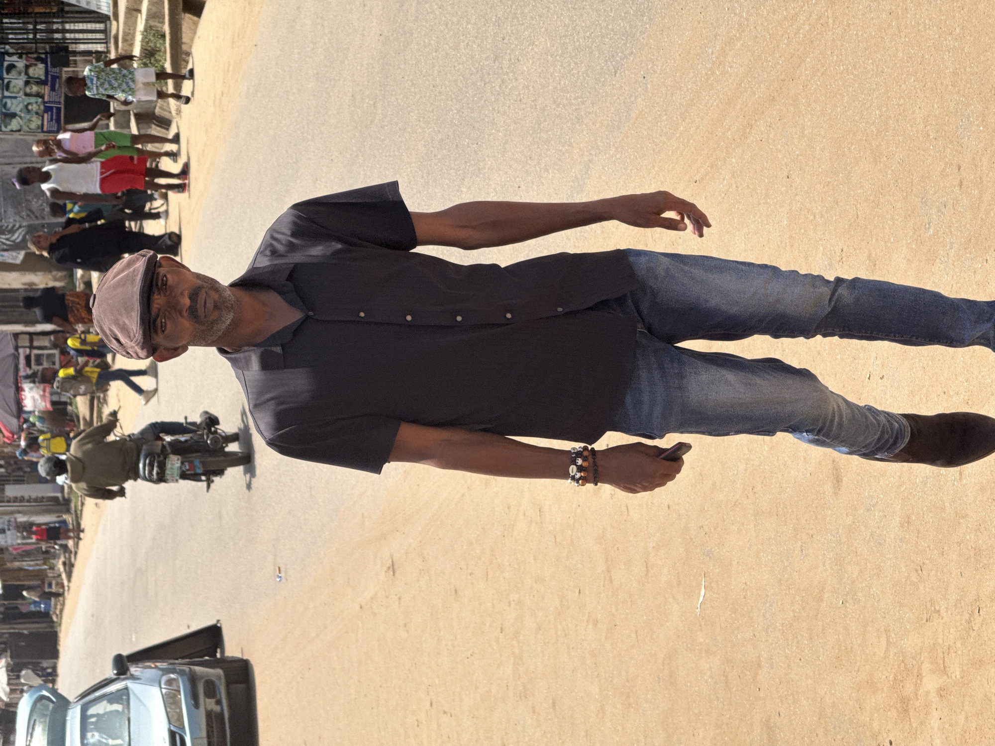 In a sunny outdoor scene on a street in Abuja, Nigeria, a man in his late 40s to early 50s with a dark complexion, wearing a dark brown flat cap, a black short-sleeved button-up shirt, and blue jeans, walks towards the camera. He has a greying beard and appears to be holding a mobile phone in his right hand. On his left wrist, he wears multiple bracelets, some appearing to be made of beads and shells.
The man is walking on a dirt and asphalt road. To his left, a light blue sedan with its trunk open is parked. In the background, several people are visible. A person is riding a motorcycle, also facing away from the camera. Other individuals, including children, are walking in the distance, some near shops and structures that line the street. The environment appears to be a bustling urban or semi-urban area, possibly a market street. The lighting suggests it is daytime, with shadows indicating bright sunlight.